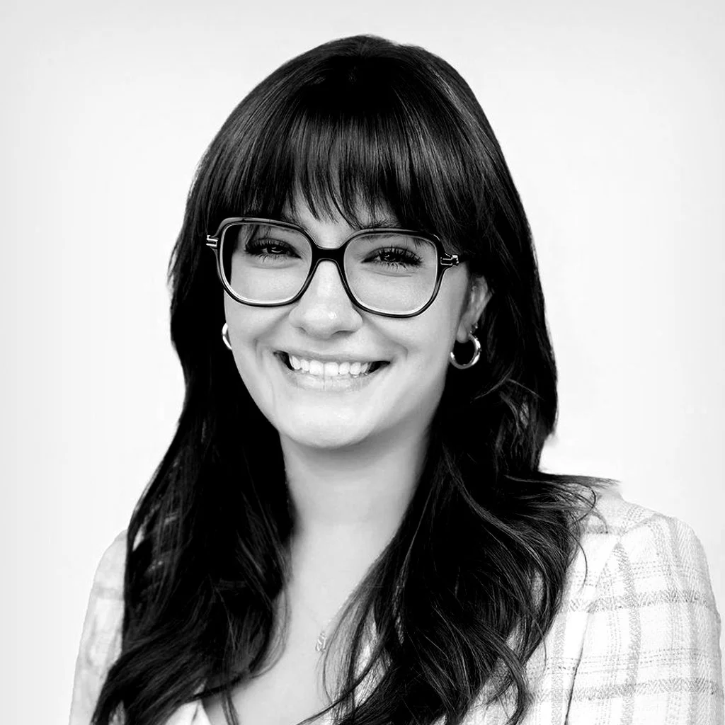 Portrait of a woman with dark hair, glasses, earrings, and a smile, against a plain background.