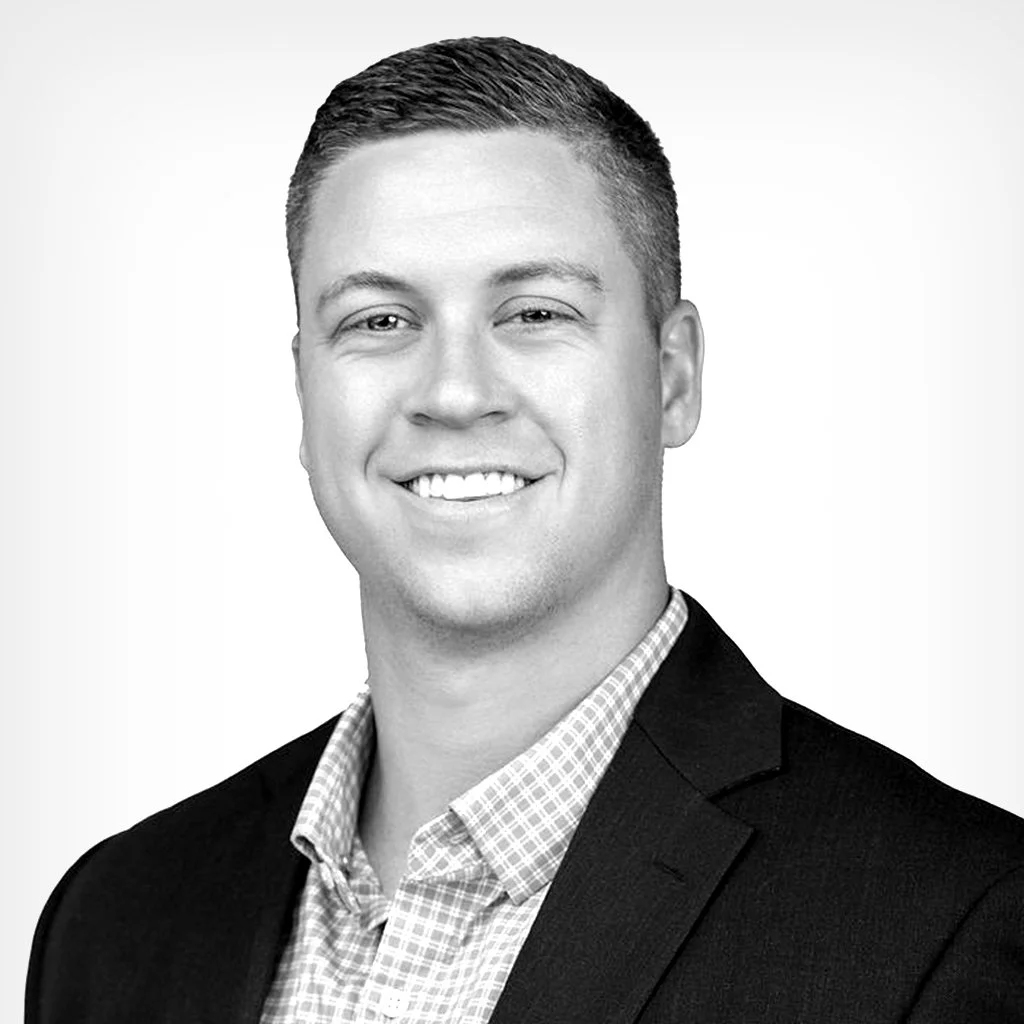 Black and white professional headshot of a smiling man wearing a suit jacket and a checkered shirt.