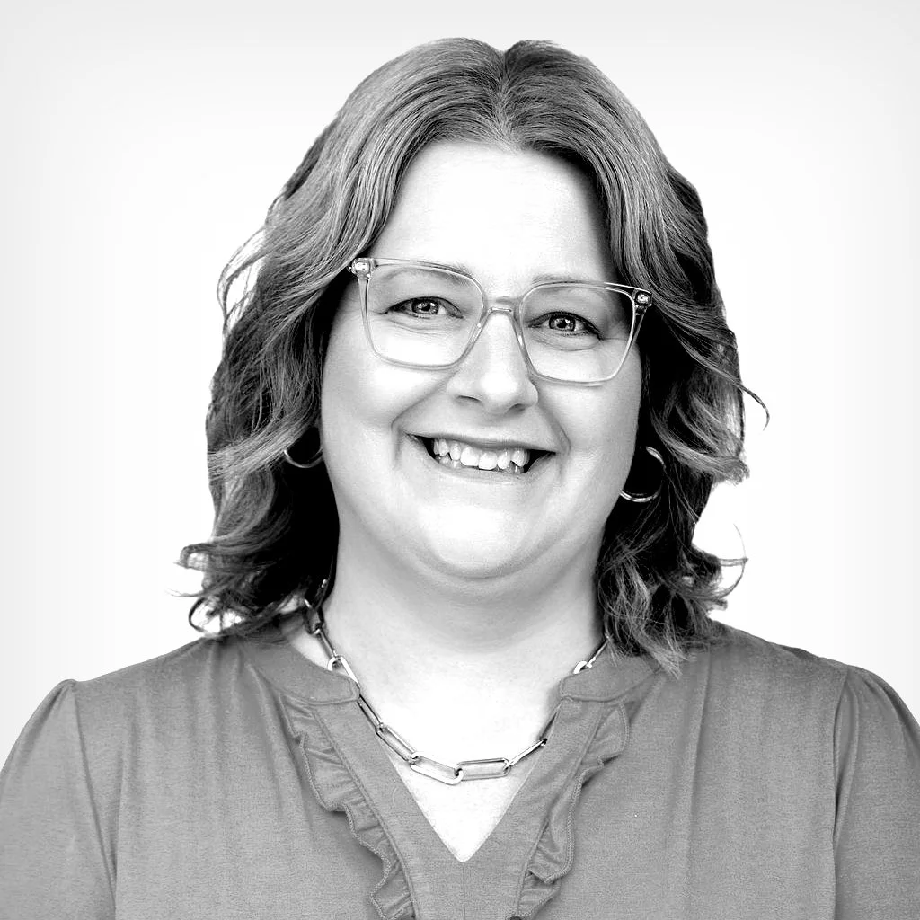 Black and white portrait of a smiling woman with glasses, shoulder-length wavy hair, wearing a blouse and a chain necklace.