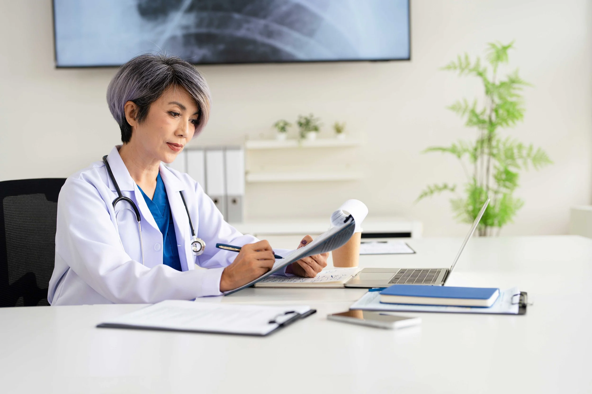 A female doctor sitting at a white desk, looking at a clipboard. She has short, grayish-purple hair and is wearing a white coat with a stethoscope around her neck. The workspace has a laptop, notebooks, and a paper cup, with a potted plant and a large television screen behind her in a medical office.