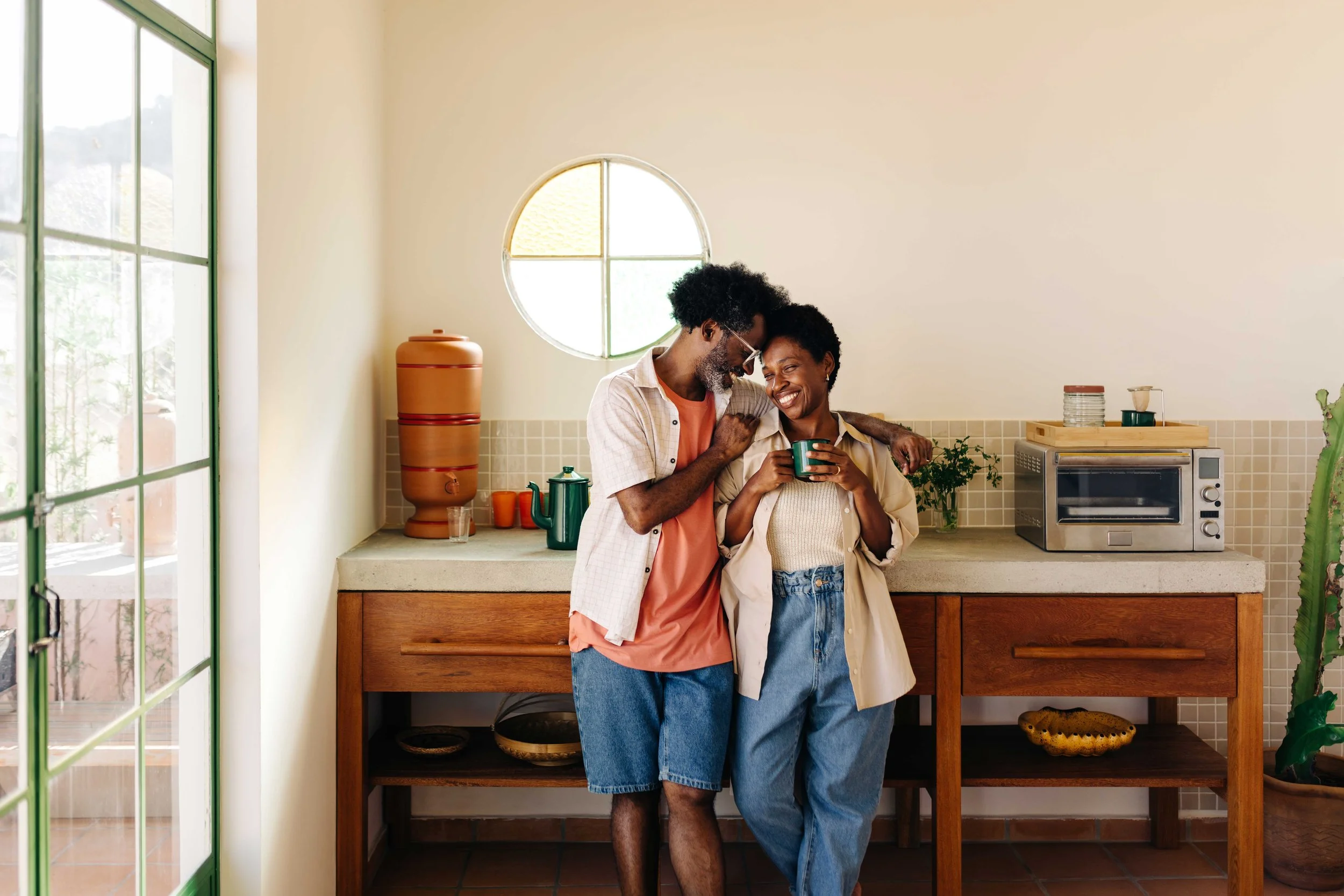 A happy couple standing close together in a kitchen, smiling and enjoying each other's company. The man has a beard and glasses, and the woman has short curly hair. The kitchen has a light beige wall with a round window and a countertop with plants, a toaster oven, and other kitchen items.