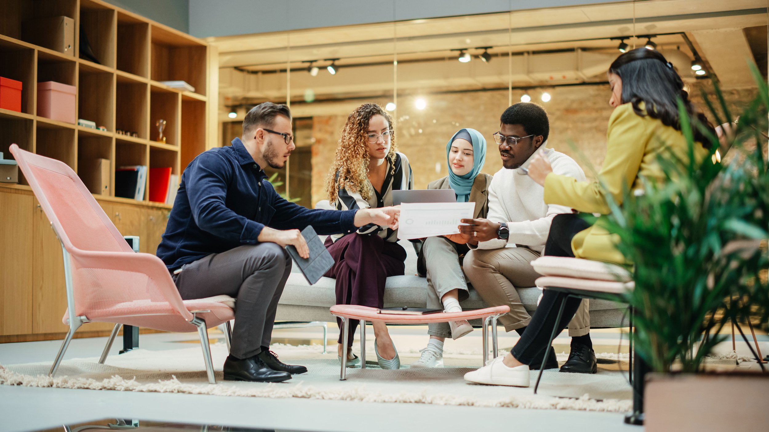 A diverse group of five people sitting closely together in a modern office lounge area, engaging in a discussion while looking at documents and a tablet, with a wooden shelf and exposed brick wall in the background.