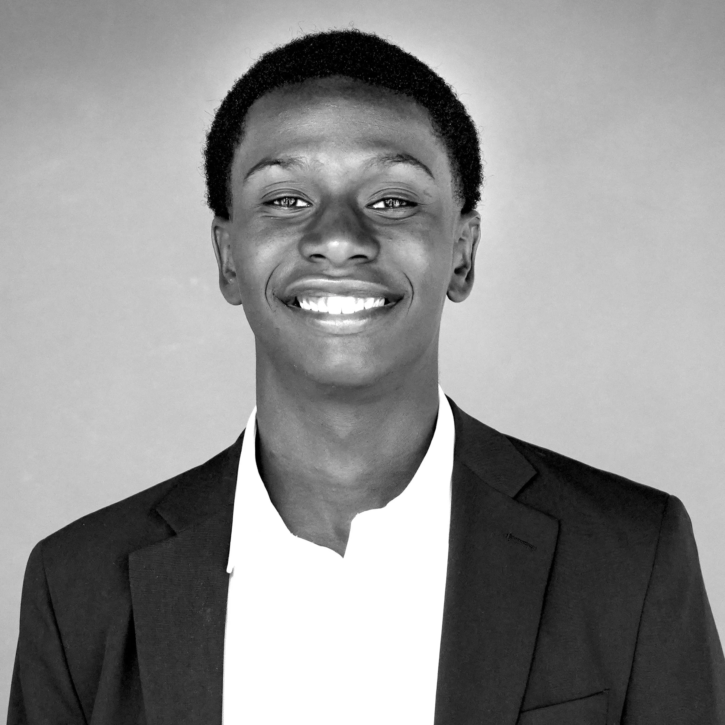 Close-up black and white photo of a person smiling, wearing a suit and tie, with short curly hair.