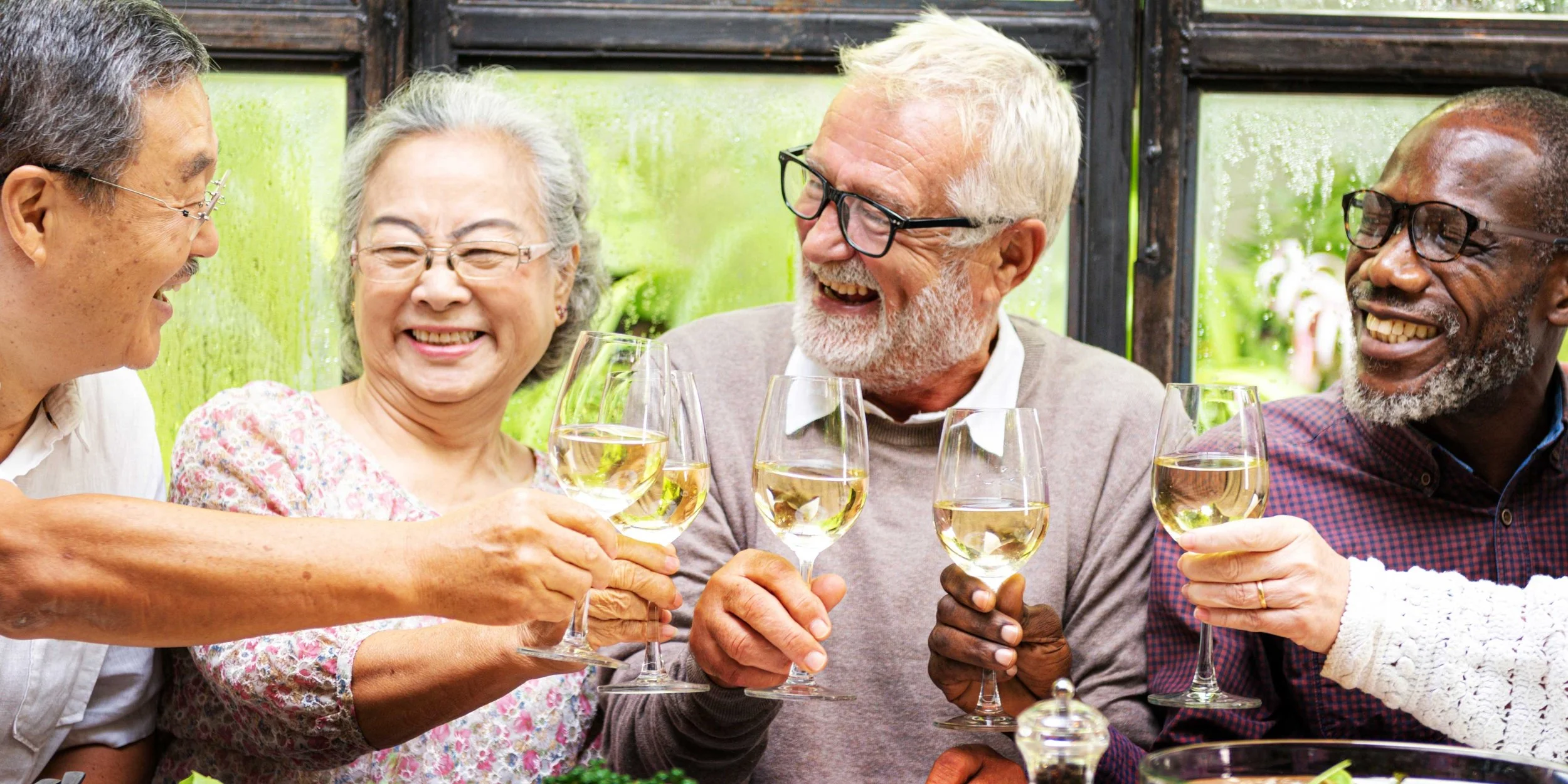 A diverse group of senior friends raising glasses of white wine and smiling at each other at an outdoor dining table.