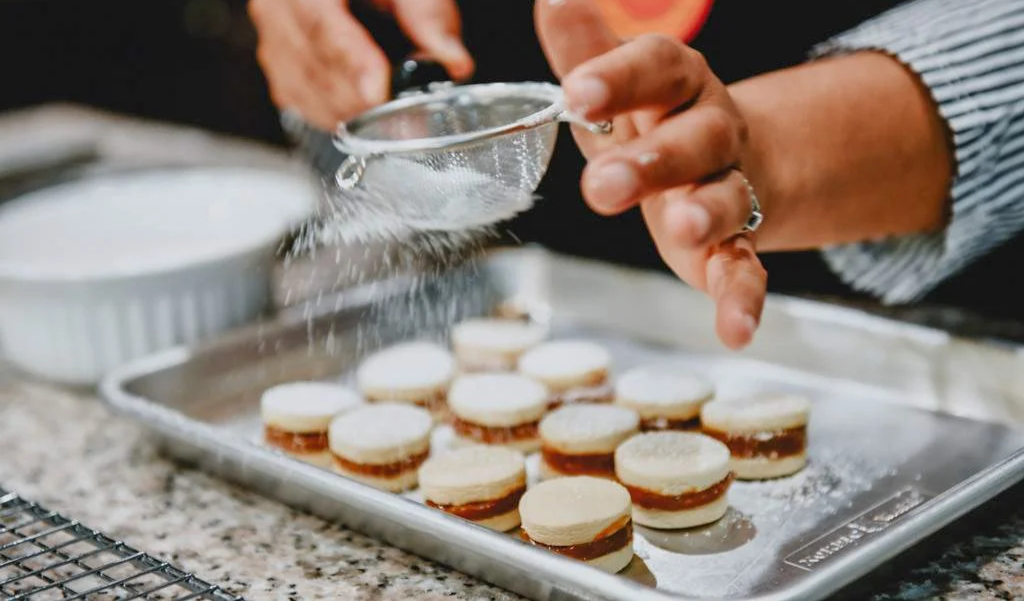 A person dusting powdered sugar over mini macarons on a baking sheet.