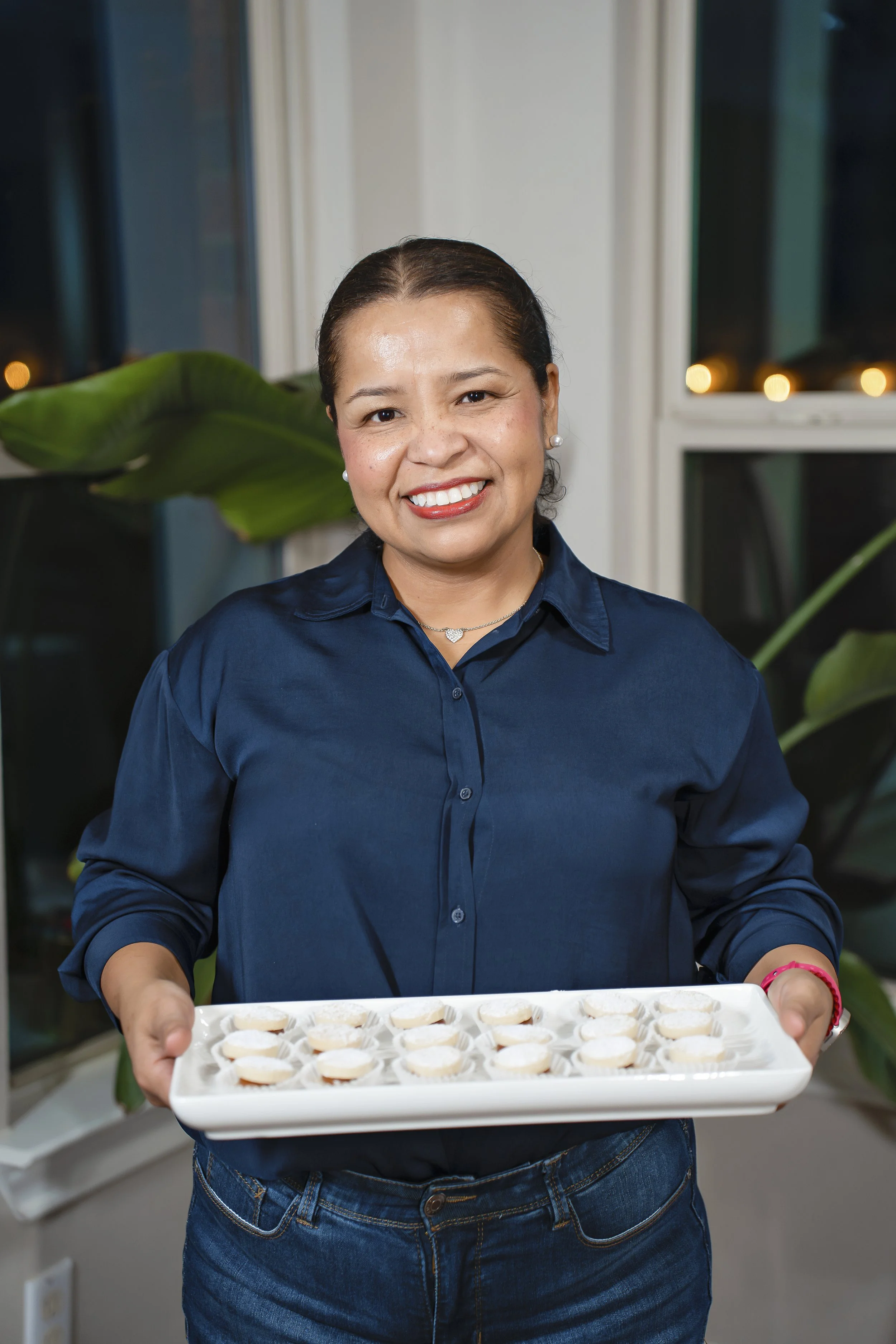 A woman smiling and holding a tray of small, round appetizers in a well-lit indoor setting with large windows and green plants in the background.