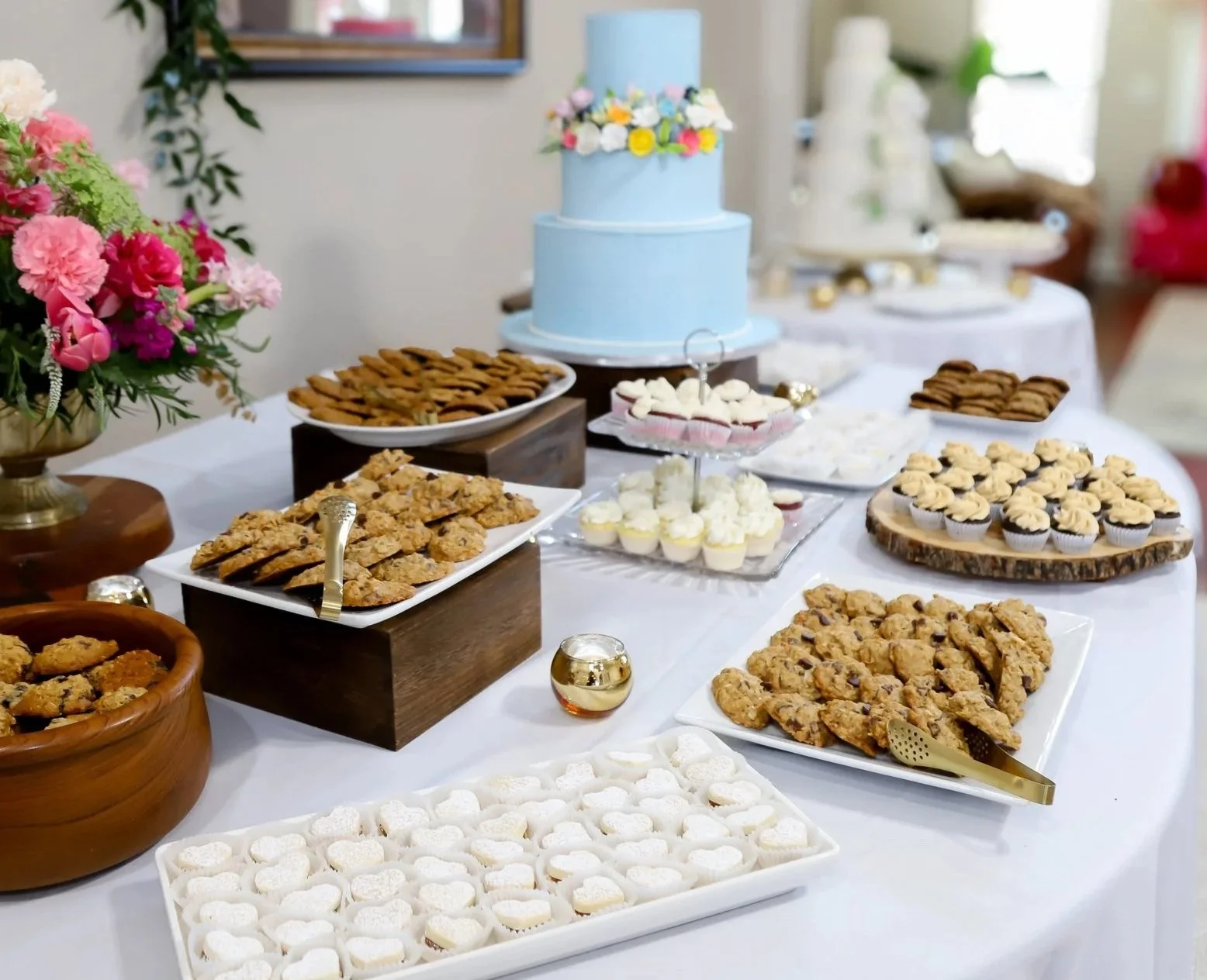 A dessert table with a light blue two-tier cake decorated with colorful flowers, surrounded by cookies, cupcakes, and candies on white trays and plates, with pink flowers on the side.