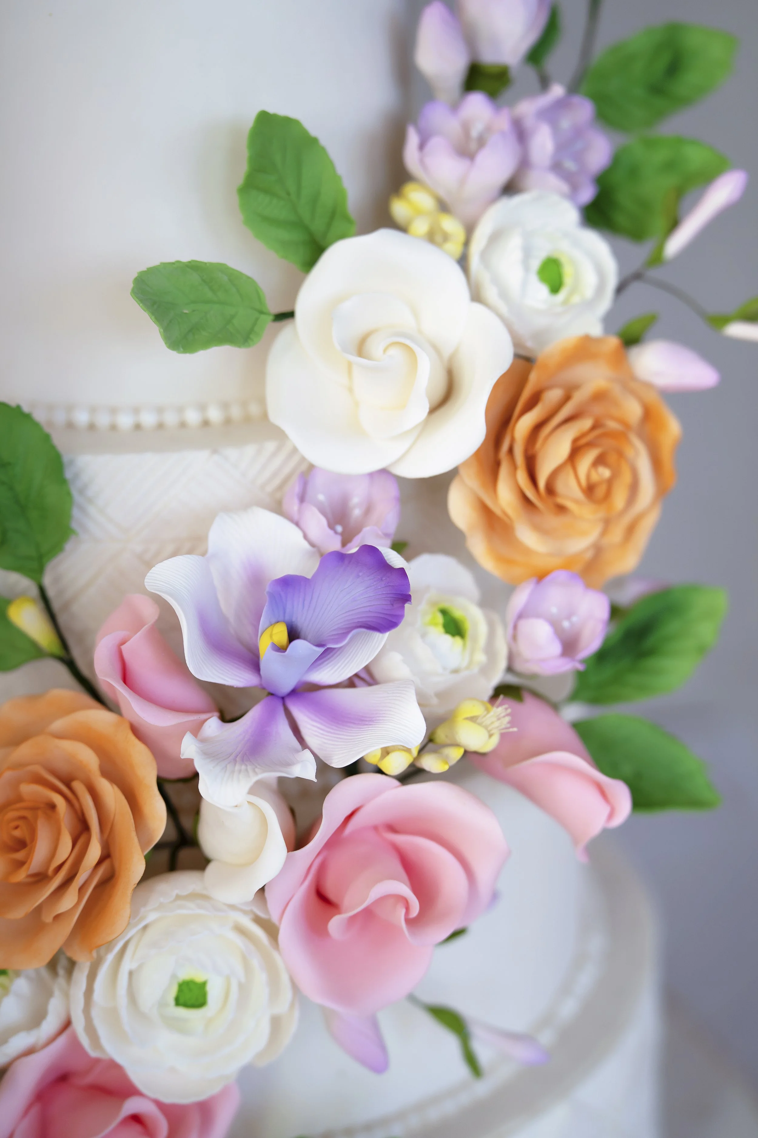 Close-up of a decorated cake with pastel-colored sugar flowers, including roses, calla lilies, and other blossoms, with green leaves as decoration.
