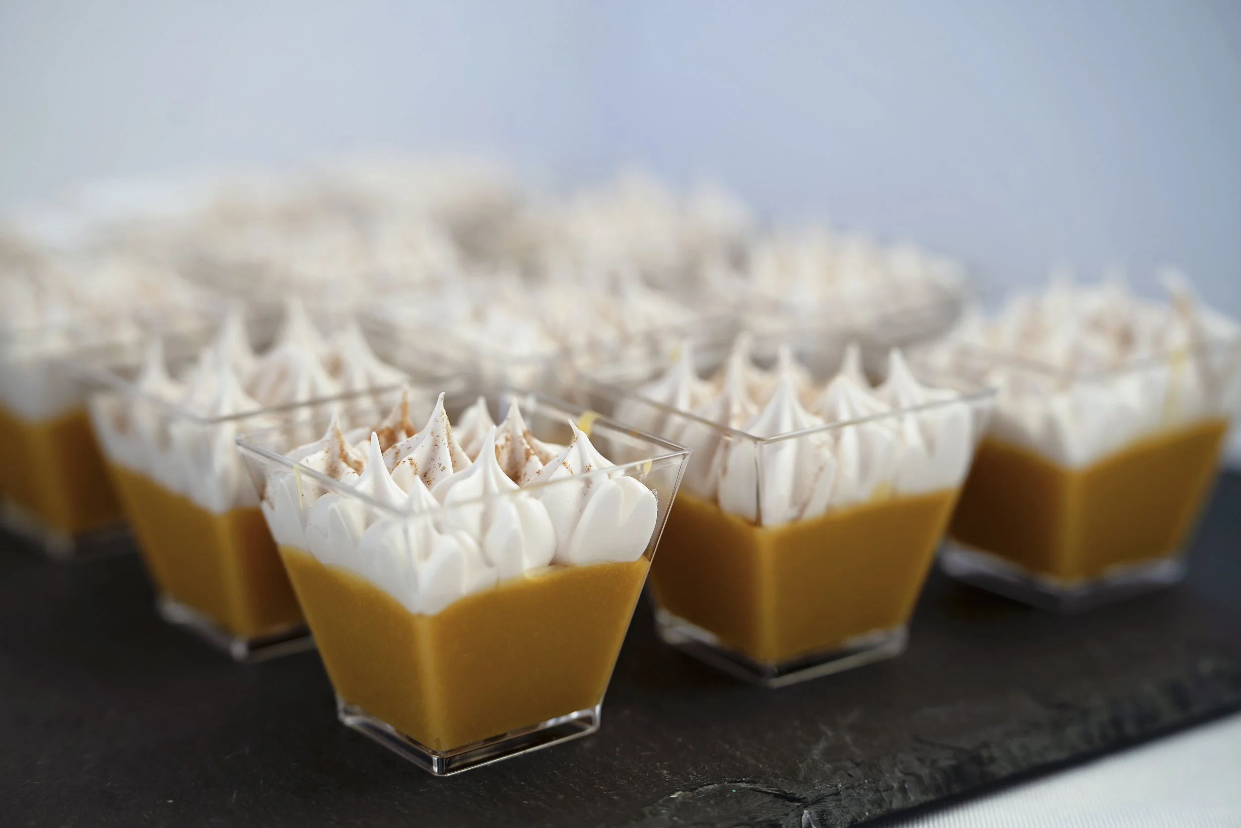 Several cups of caramel pudding topped with whipped cream and sprinkled with cinnamon, arranged on a black serving tray.