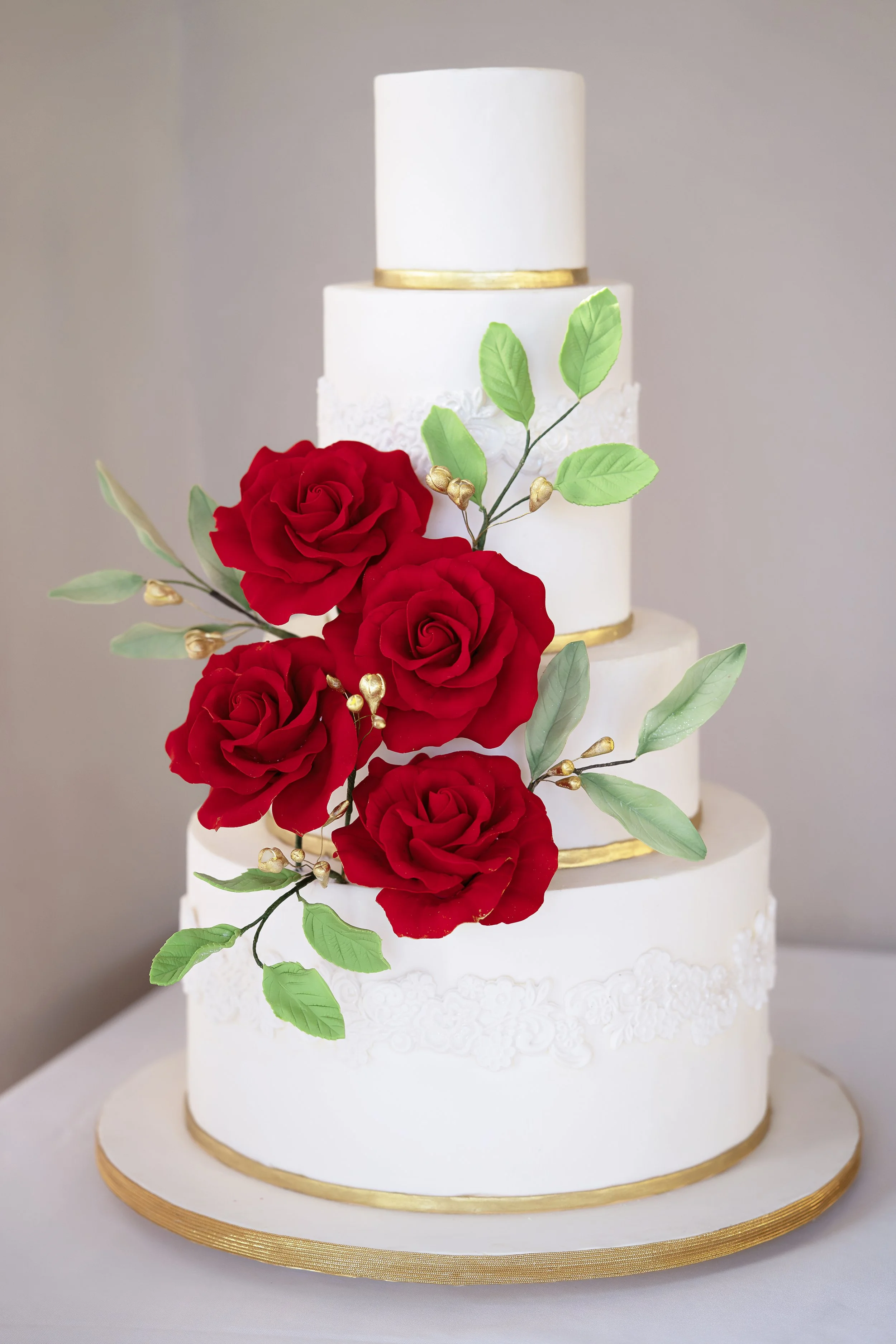 A tiered white wedding cake decorated with red roses, green leaves, and gold accents.