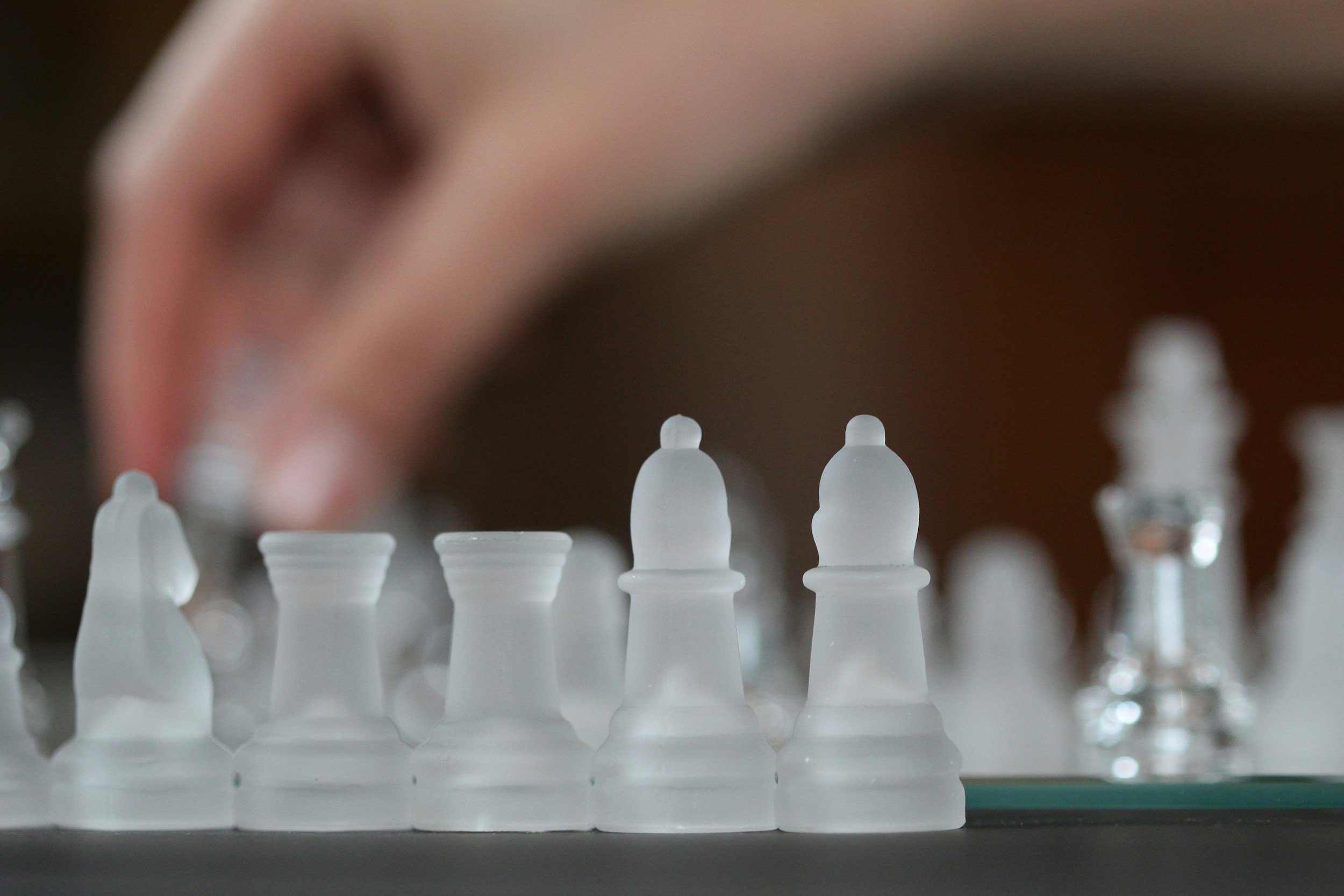 Close-up of a chessboard with frosted glass chess pieces, focusing on the pawn and rook in the foreground, with a person's hand moving a chess piece in the background.
