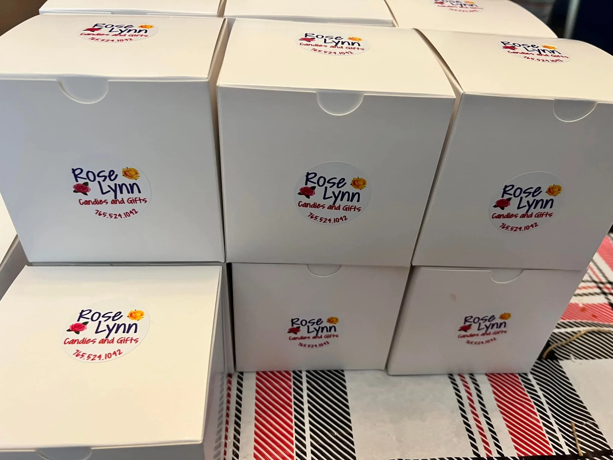 White boxes with 'Rose Lynn Candies and Gifts' logo, stacked on a table with red, black, and white plaid tablecloth.