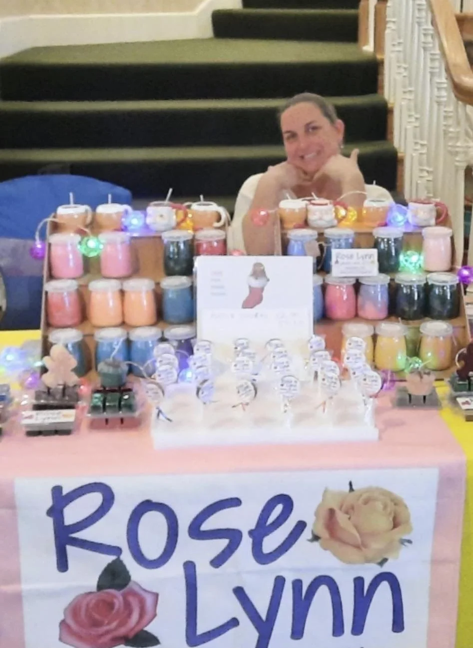 A woman sitting behind a table selling jars of homemade jams, with a hand-painted sign that says "Rose Lynn" and pictures of roses, decorated with colorful string lights.