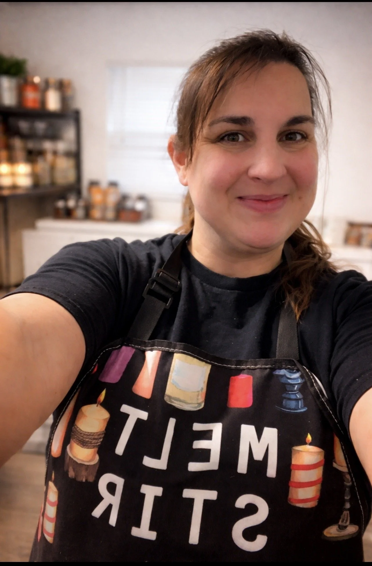 A woman taking a selfie in a kitchen, wearing a black apron with birthday cakes and candles, with a shelf of jars behind her.