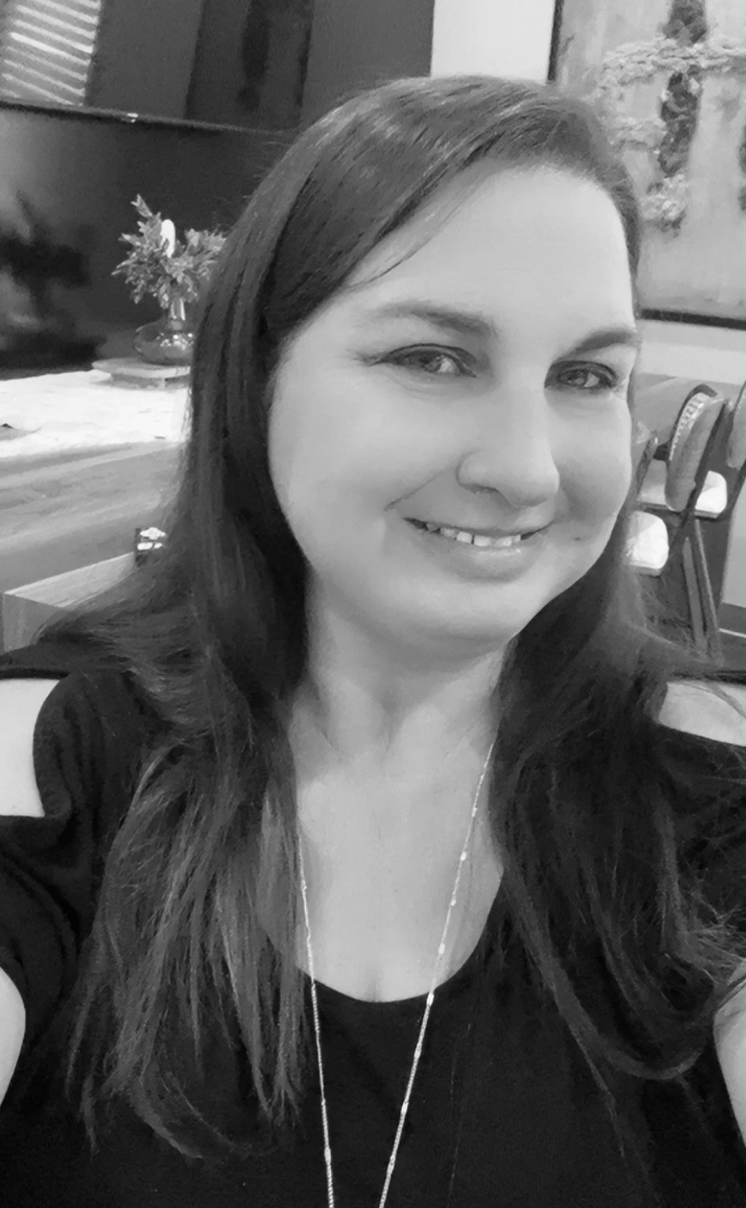 A woman with long dark hair smiling for a selfie in a room with decorated walls and a table with a flower arrangement in the background.