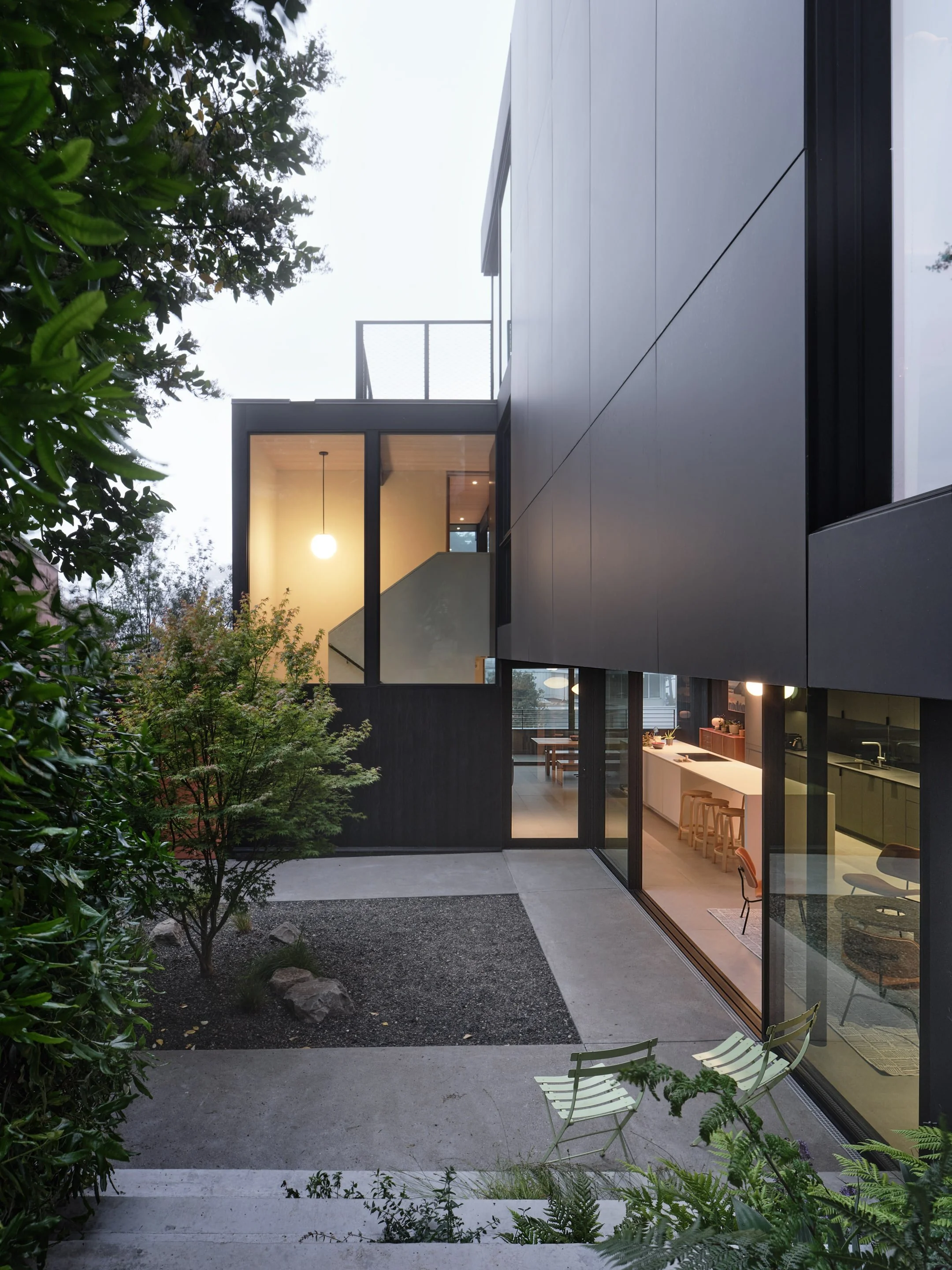 Modern multi-story house with large glass windows, black exterior panels, and an outdoor courtyard with seating, greenery, and stairs.