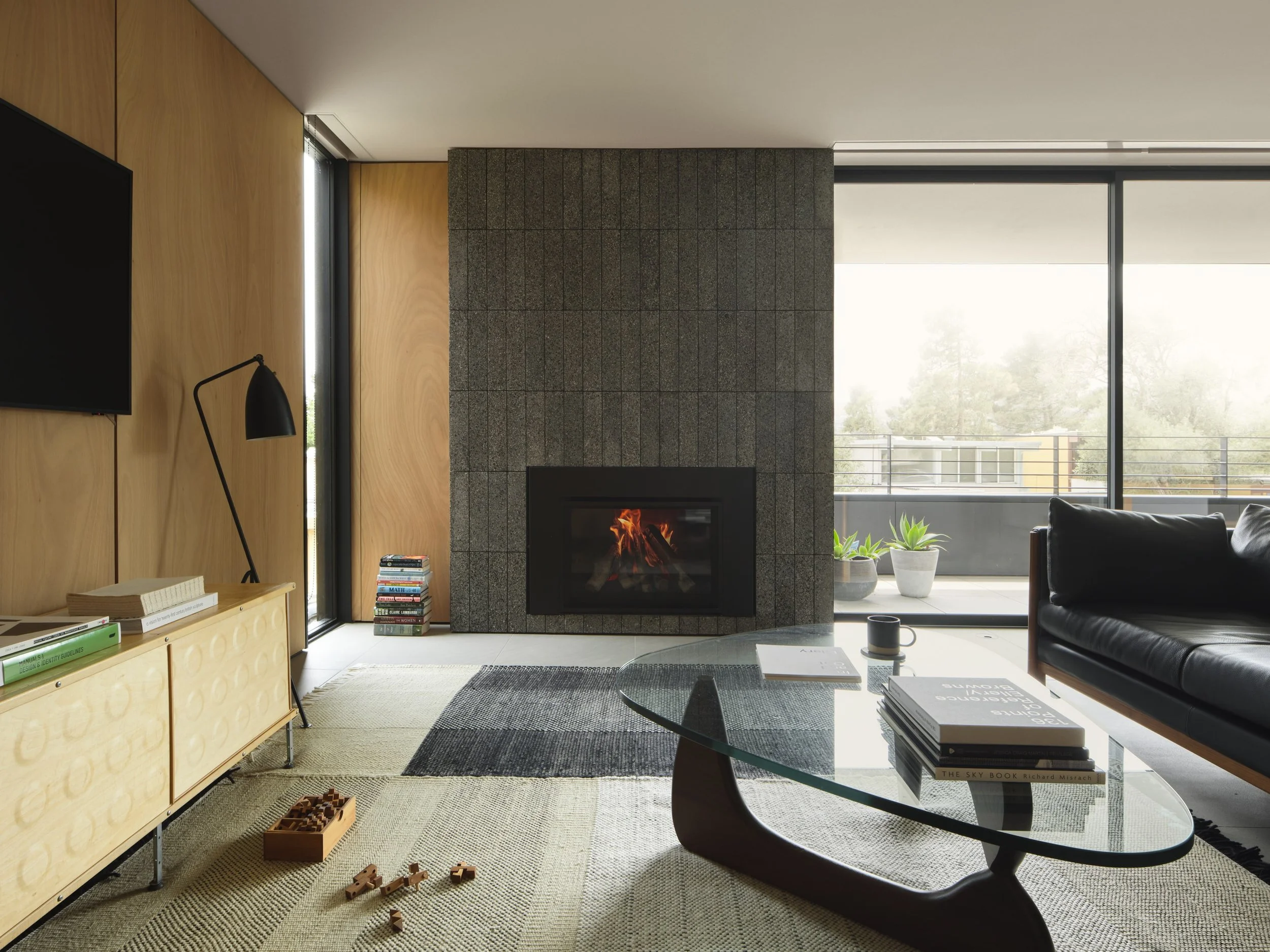 Modern living room with a central fireplace, black leather sofa, glass coffee table, books, and a large sliding glass door leading to a balcony with potted plants.