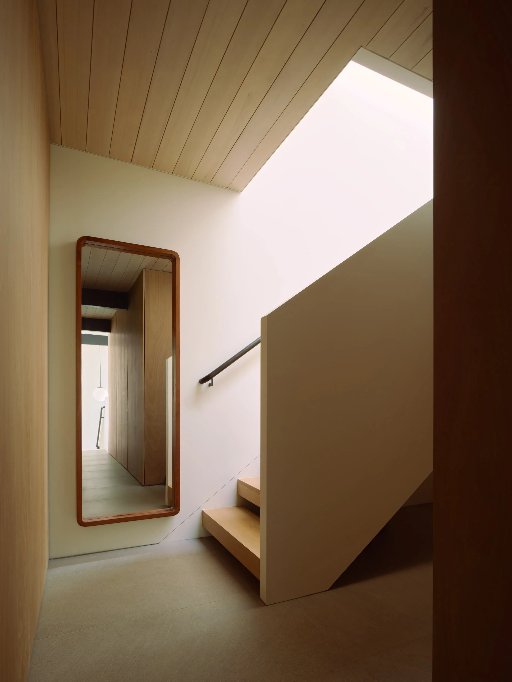 Modern interior staircase with wooden steps, white side panel, black handrail, wooden ceiling, a tall mirror with rounded wooden frame, and natural light from a large skylight.