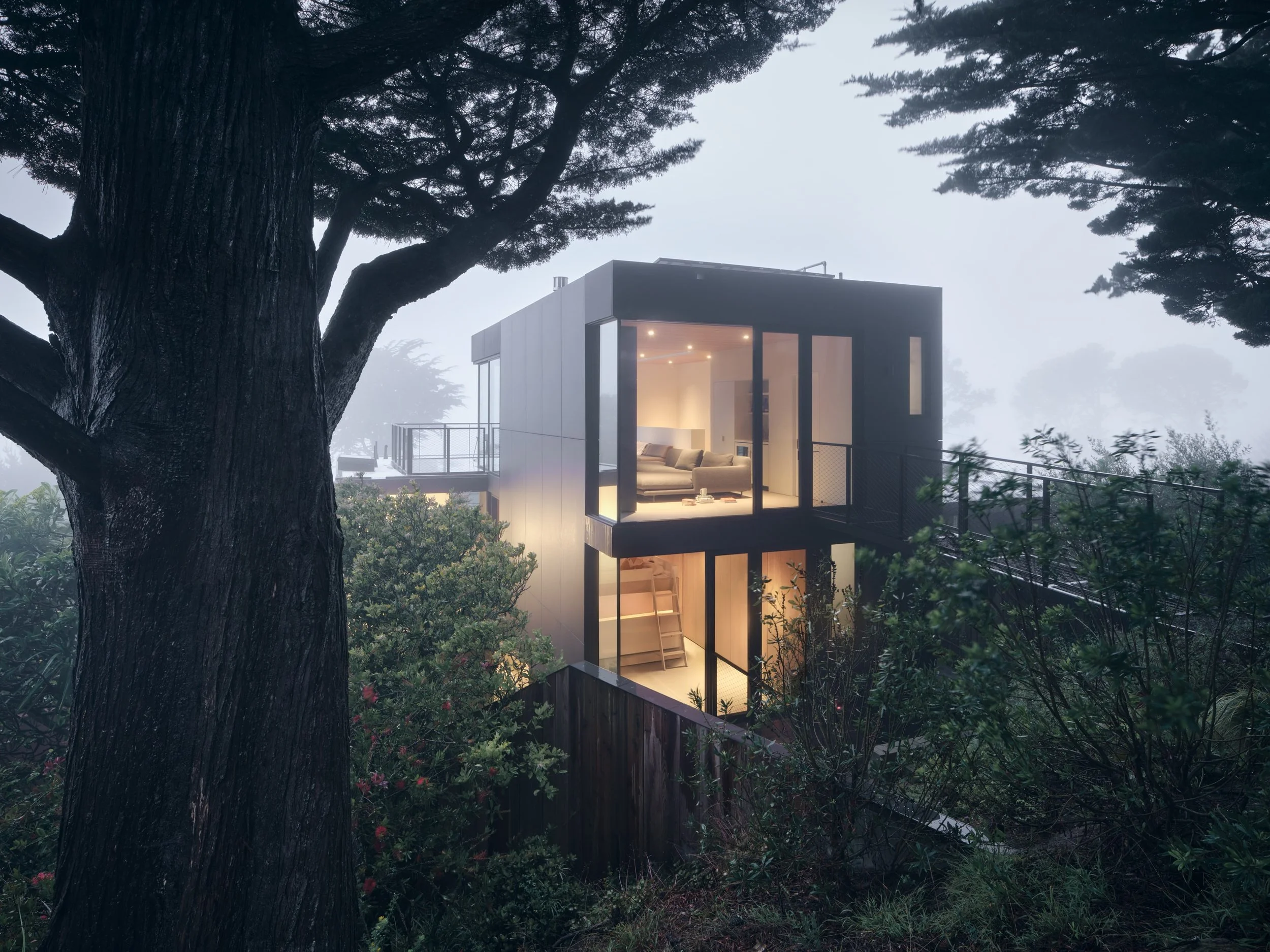 Modern two-story house nestled among trees with large glass windows, illuminated from within, on a foggy day.