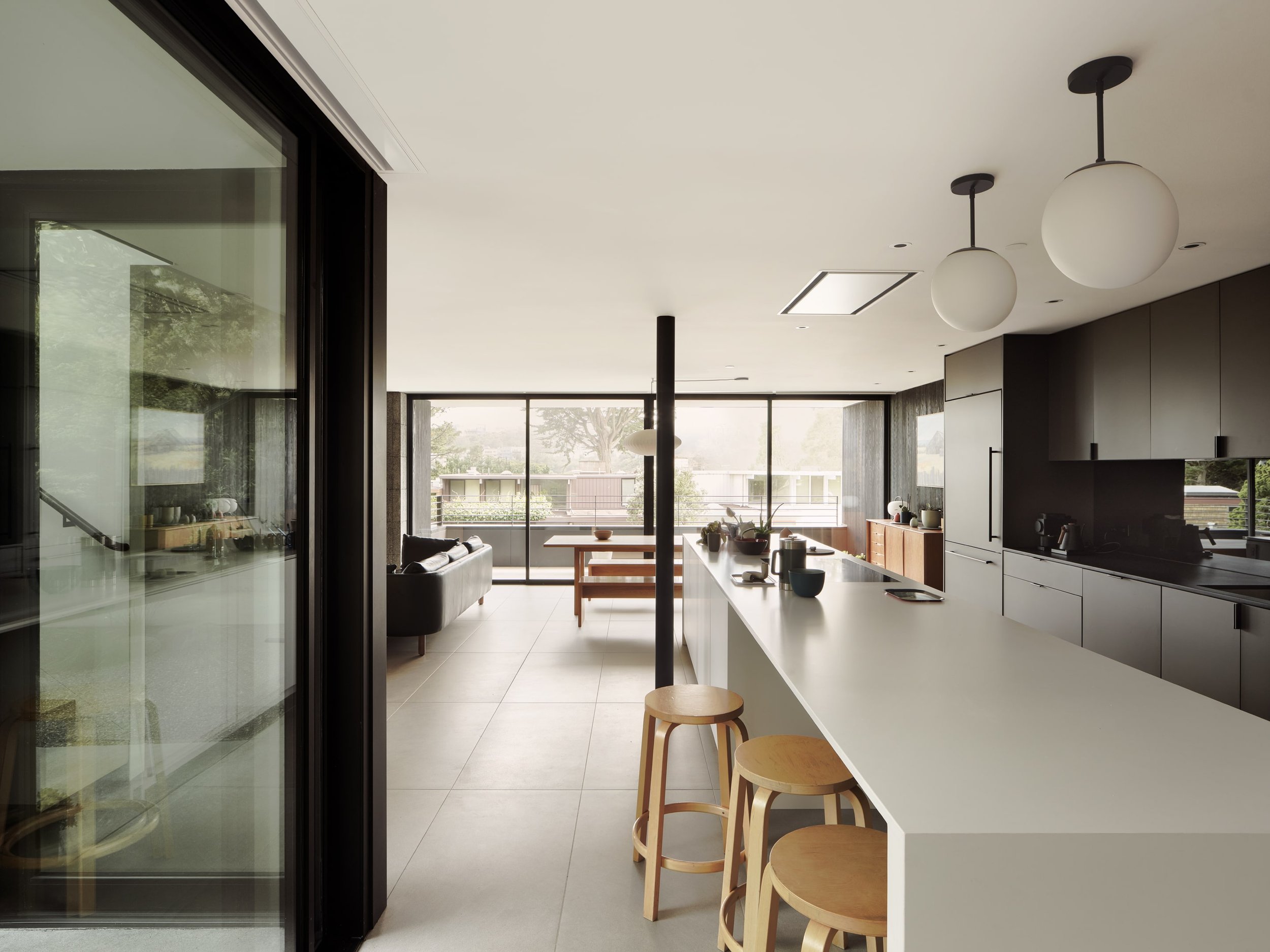 Modern kitchen with white island, black cabinets, three round white pendant lights, and large windows overlooking outdoor greenery.