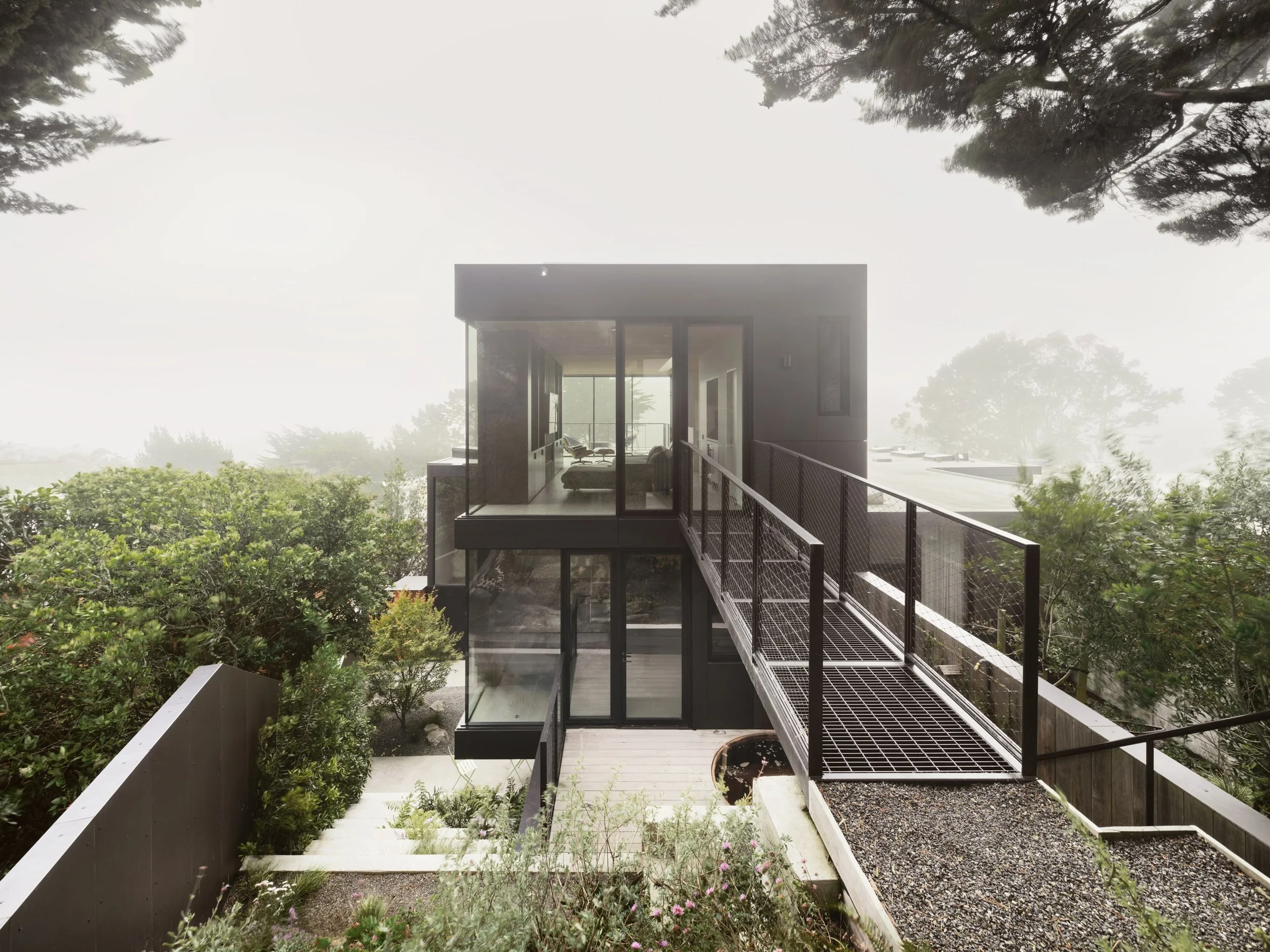 Modern two-story house with large glass windows, a rooftop deck, and a metal walkway railing, surrounded by trees and greenery.