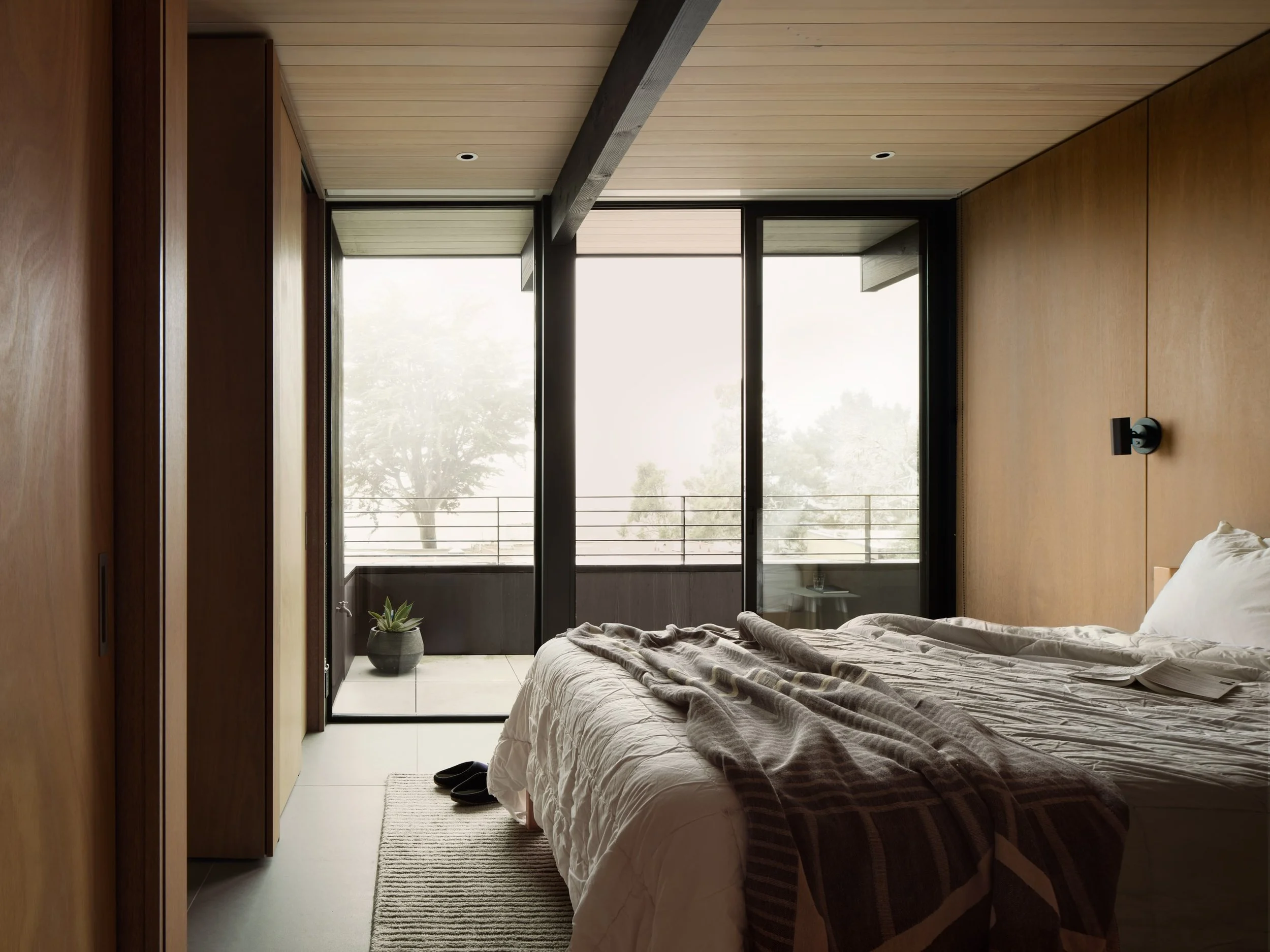 Modern bedroom with wooden walls, large sliding glass doors leading to a balcony, a bed with white bedding and a plaid blanket, a small potted plant outside, and a pair of black slippers on a rug.
