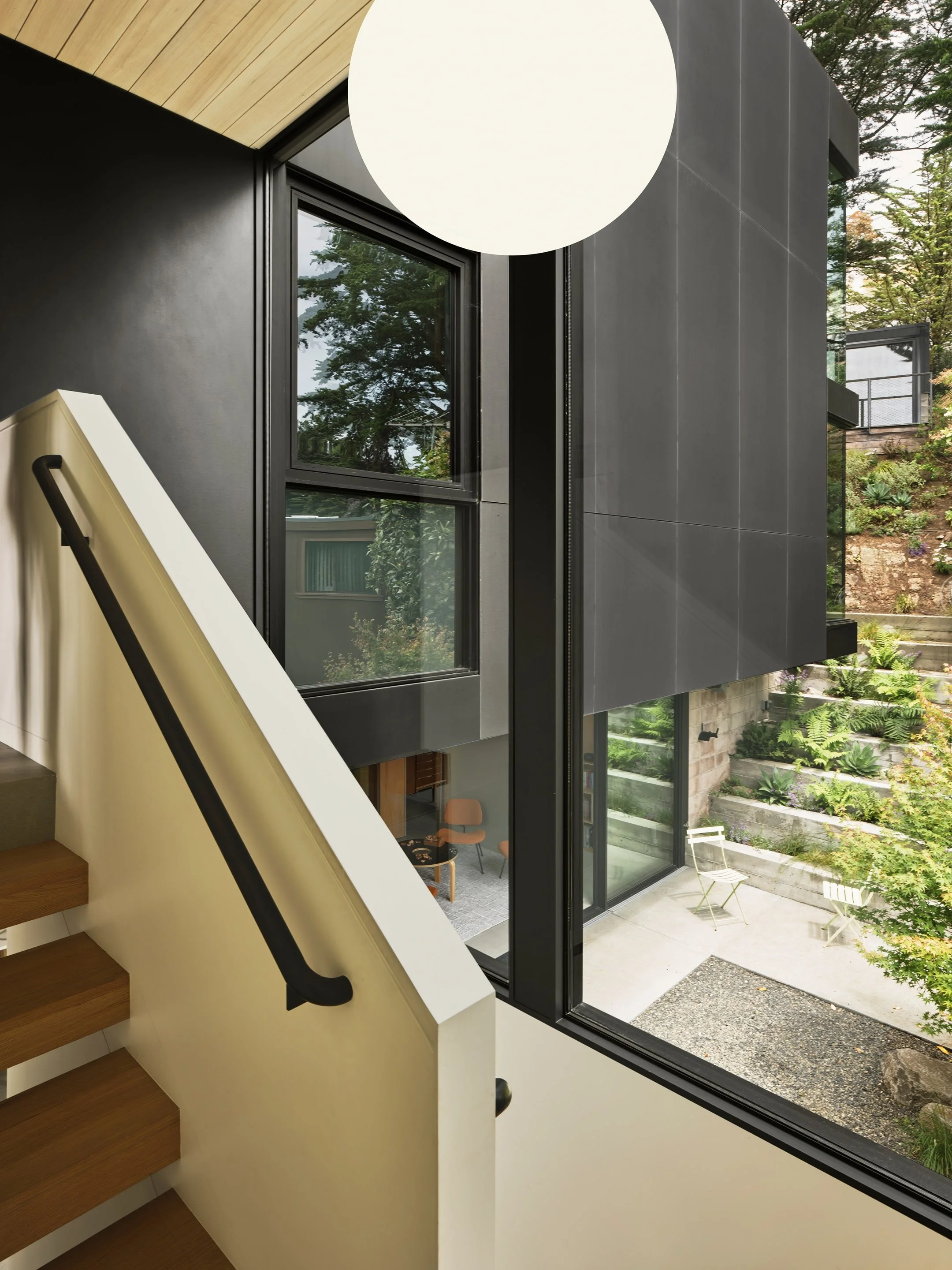 Interior view of a modern house showcasing a staircase with wooden steps and a black handrail, large windows revealing an outdoor garden, and a round ceiling light.
