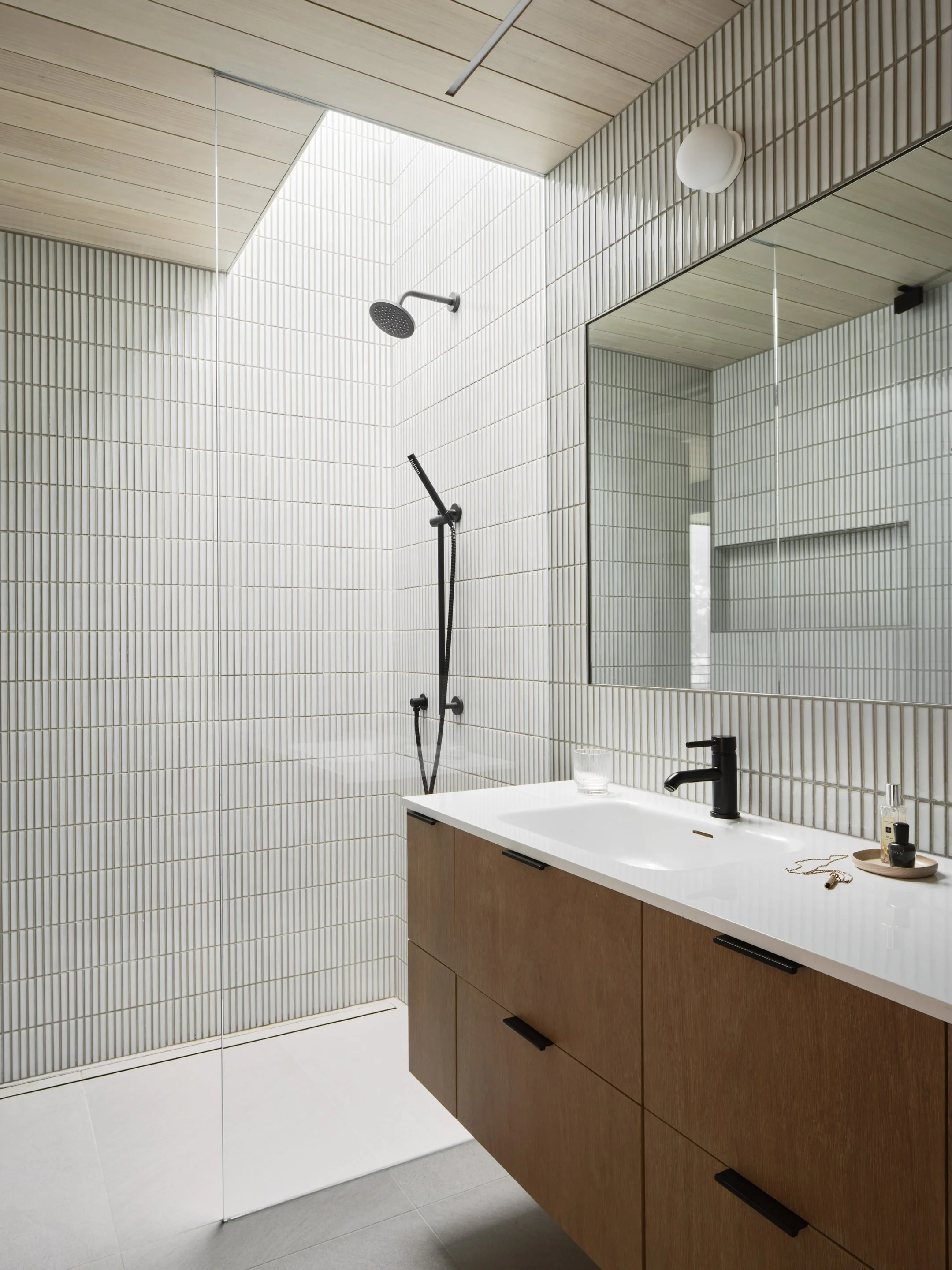 Modern minimalist bathroom with a walk-in shower on the left, a large mirror above a light-colored wooden vanity with a black faucet on the right, and white tiled walls and ceiling.