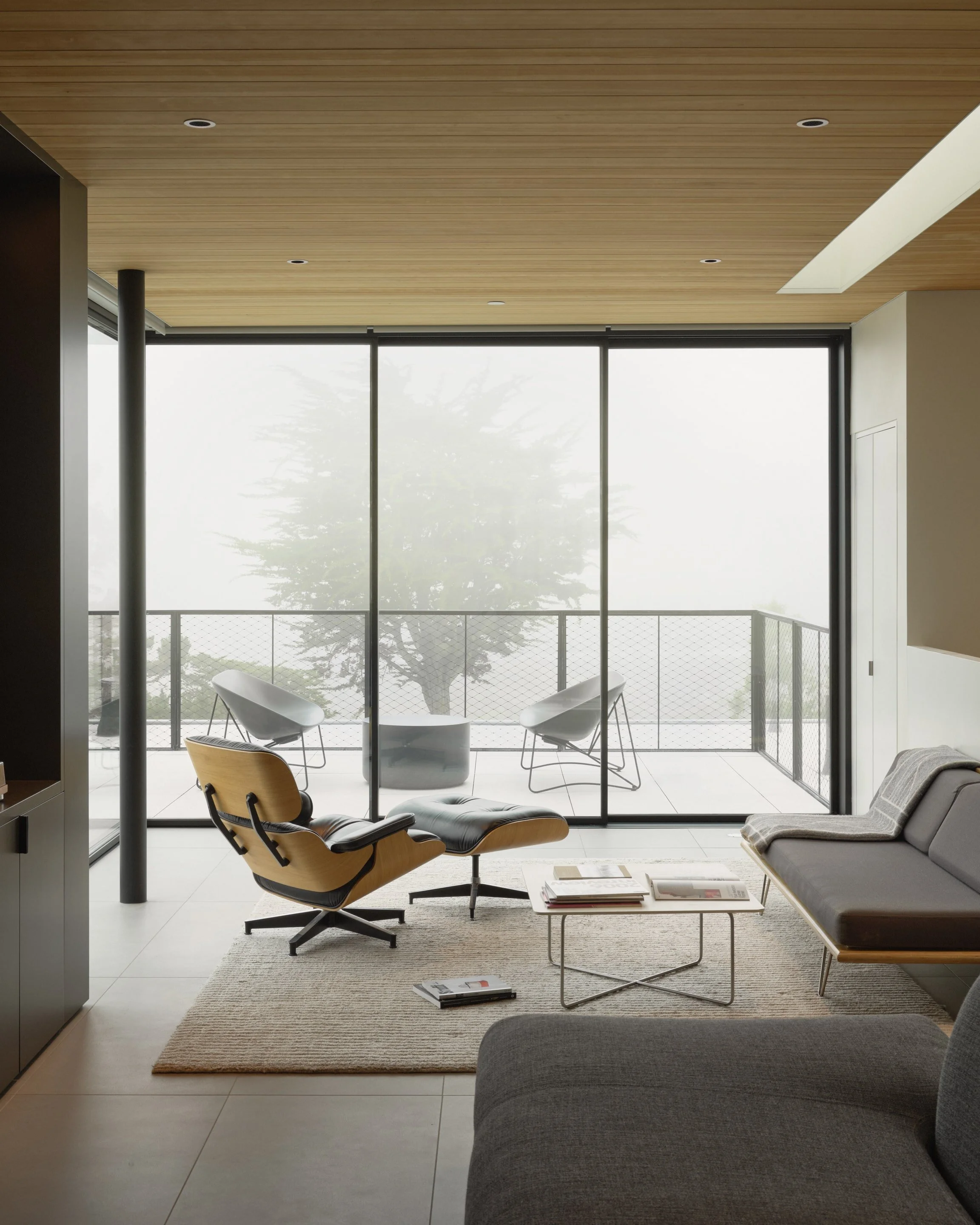 Modern living room with large glass sliding door leading to a balcony with outdoor chairs and a tree visible through foggy weather.