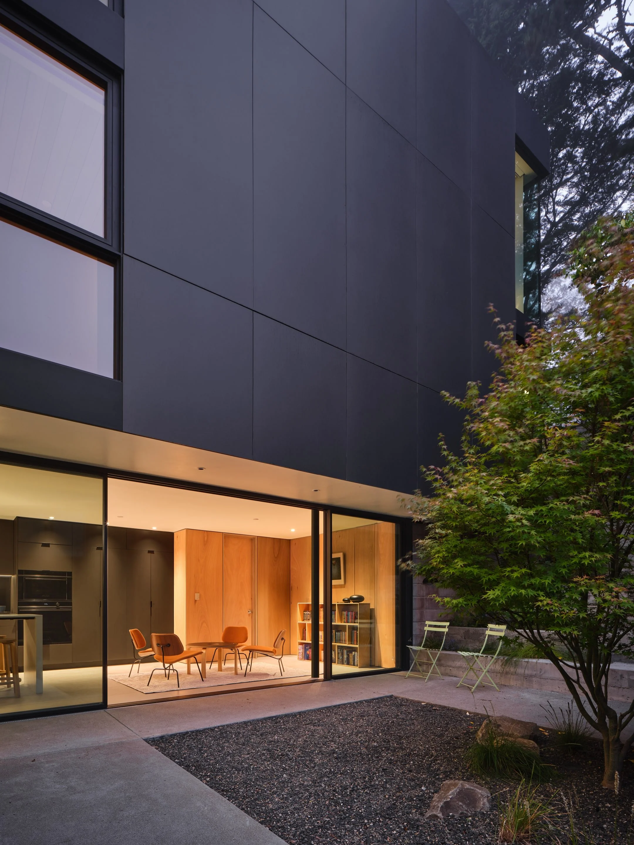 Modern house with large glass windows, showing a cozy interior with a round wooden table and orange chairs, and an outside patio with two chairs and a tree.