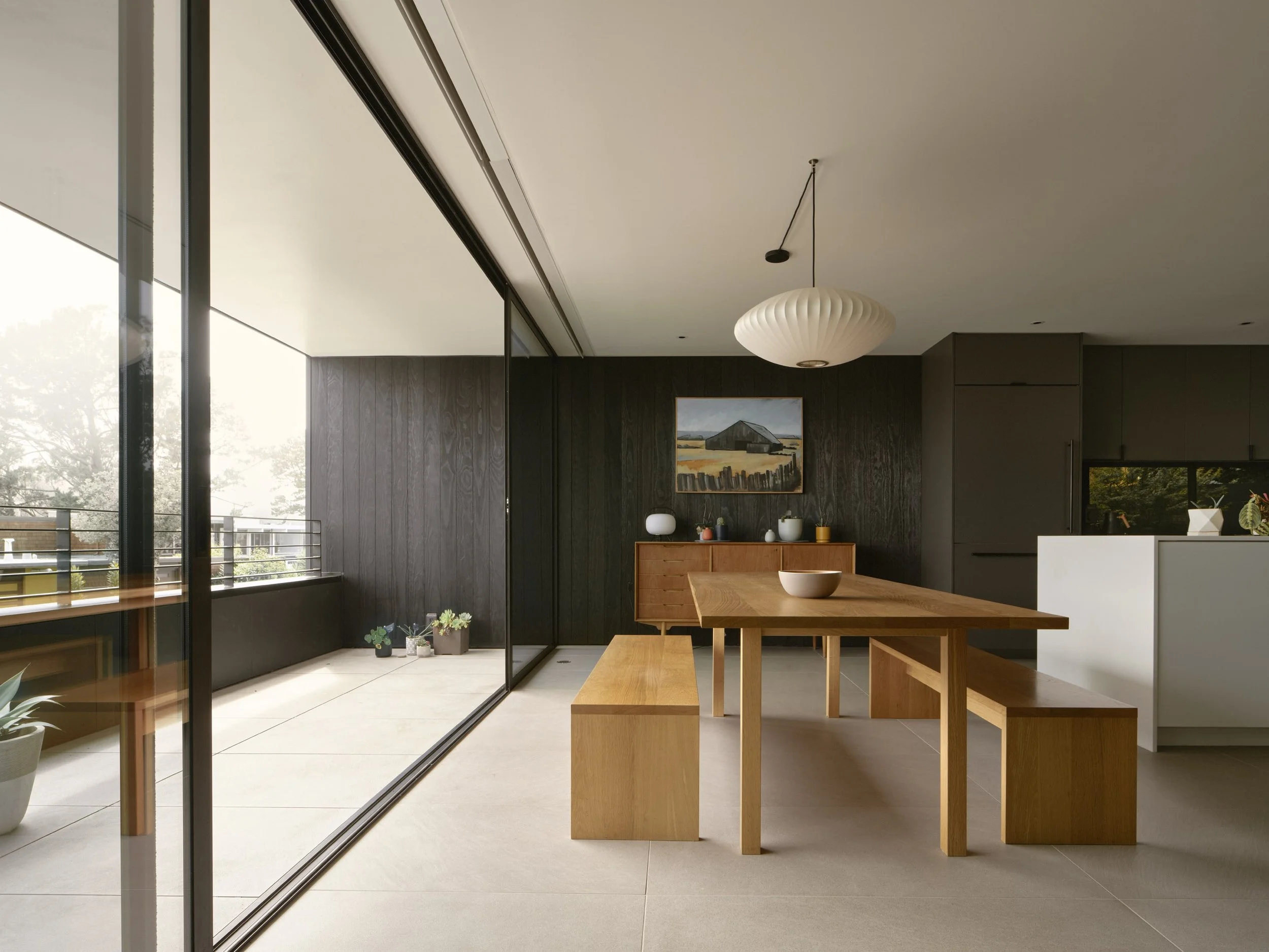 Modern dining area with wooden table and benches, large sliding glass door leading to a balcony, black accent wall with a landscape painting, white ceiling light, and minimal decor.