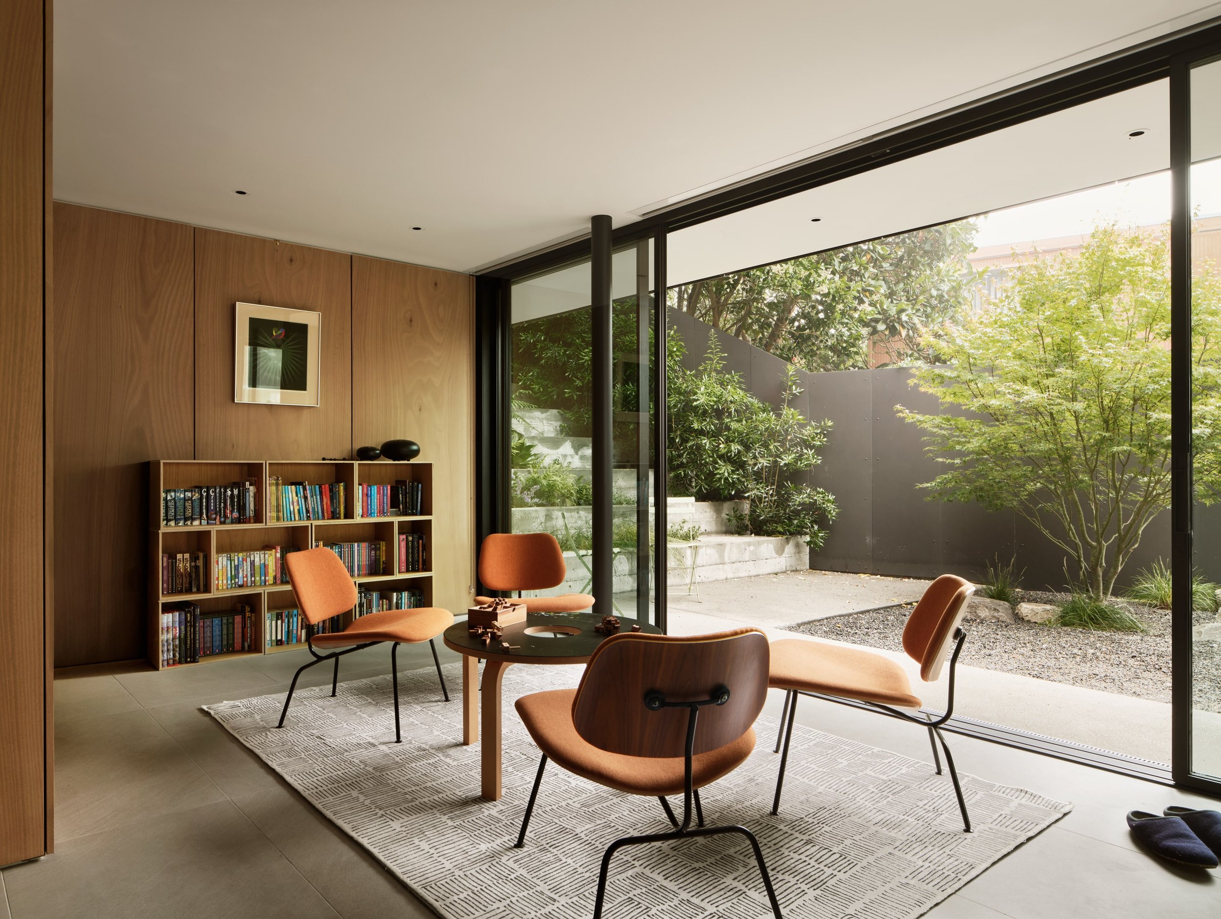 Modern living room with wooden wall panels, a bookshelf filled with books, four orange chairs around a black table, large floor-to-ceiling glass sliding doors opening to a small garden with greenery and trees.