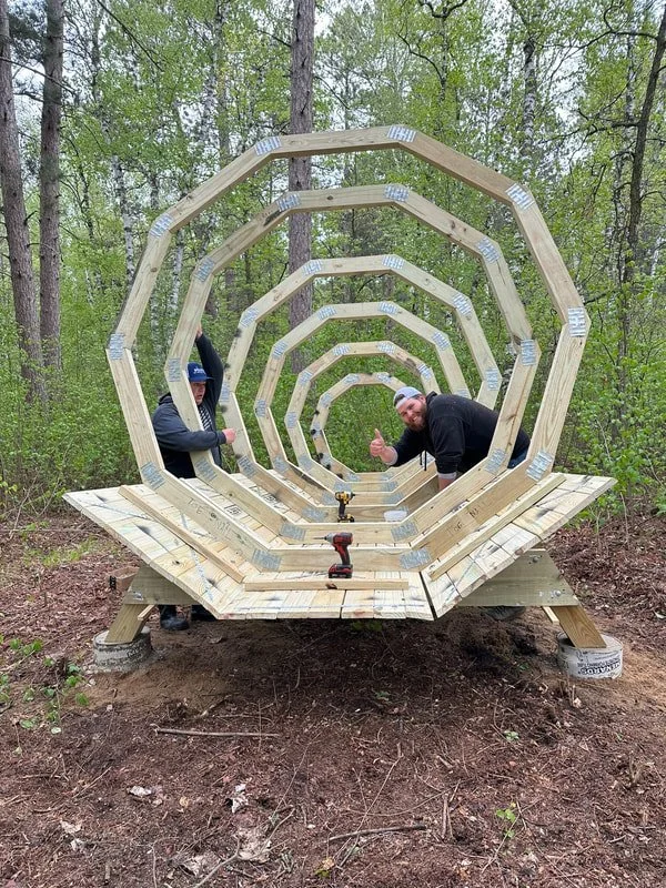Two people building a wooden shell structure for the Forest Megaphone in a wooded outdoor area. The structure consists of multiple hexagonal frames stacked and aligned, with a wooden platform base. Tools are visible on the platform.