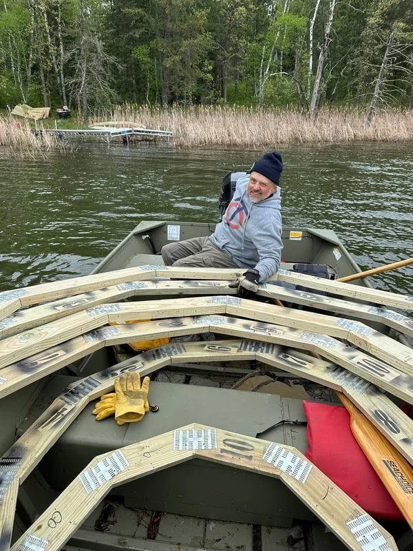 A man smiling and sitting in a boat on a lake, wearing a gray hoodie and black beanie, with wooden frames and tools in the boat, and a wooded shoreline with a dock in the background.