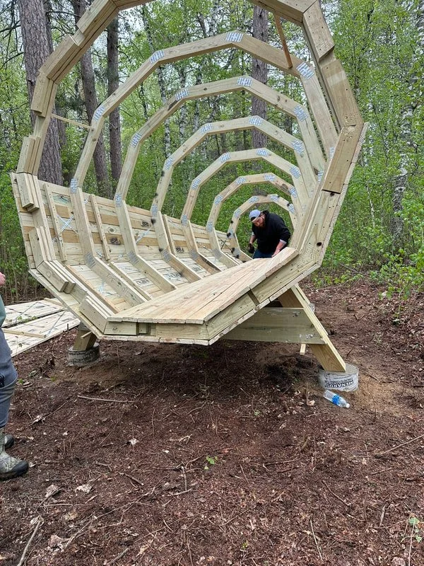 Person building a large wooden shell-shaped structure know as the Forest Megaphone outdoors in a wooded area.