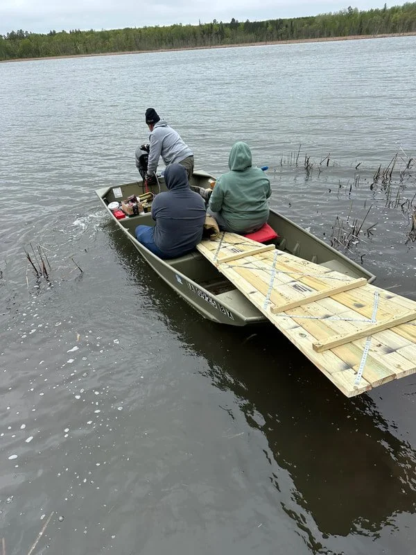 Three people in a boat with a wooden planks protruding from the front, floating on a body of water (Spearhead lake) with trees in the background.