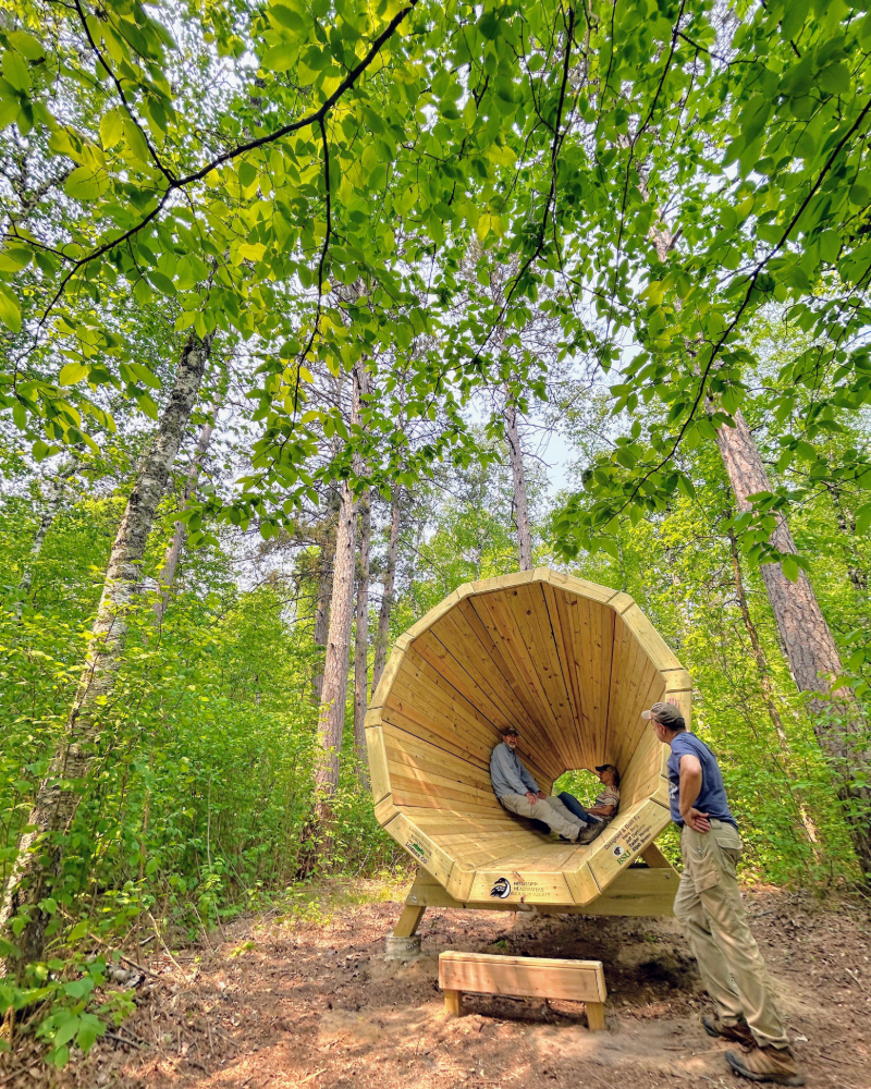 Two people sitting inside a large, wooden, octagonal outdoor structure in a forest, with another person standing outside nearby. The structure is built among green trees and has a small wooden bench underneath.
