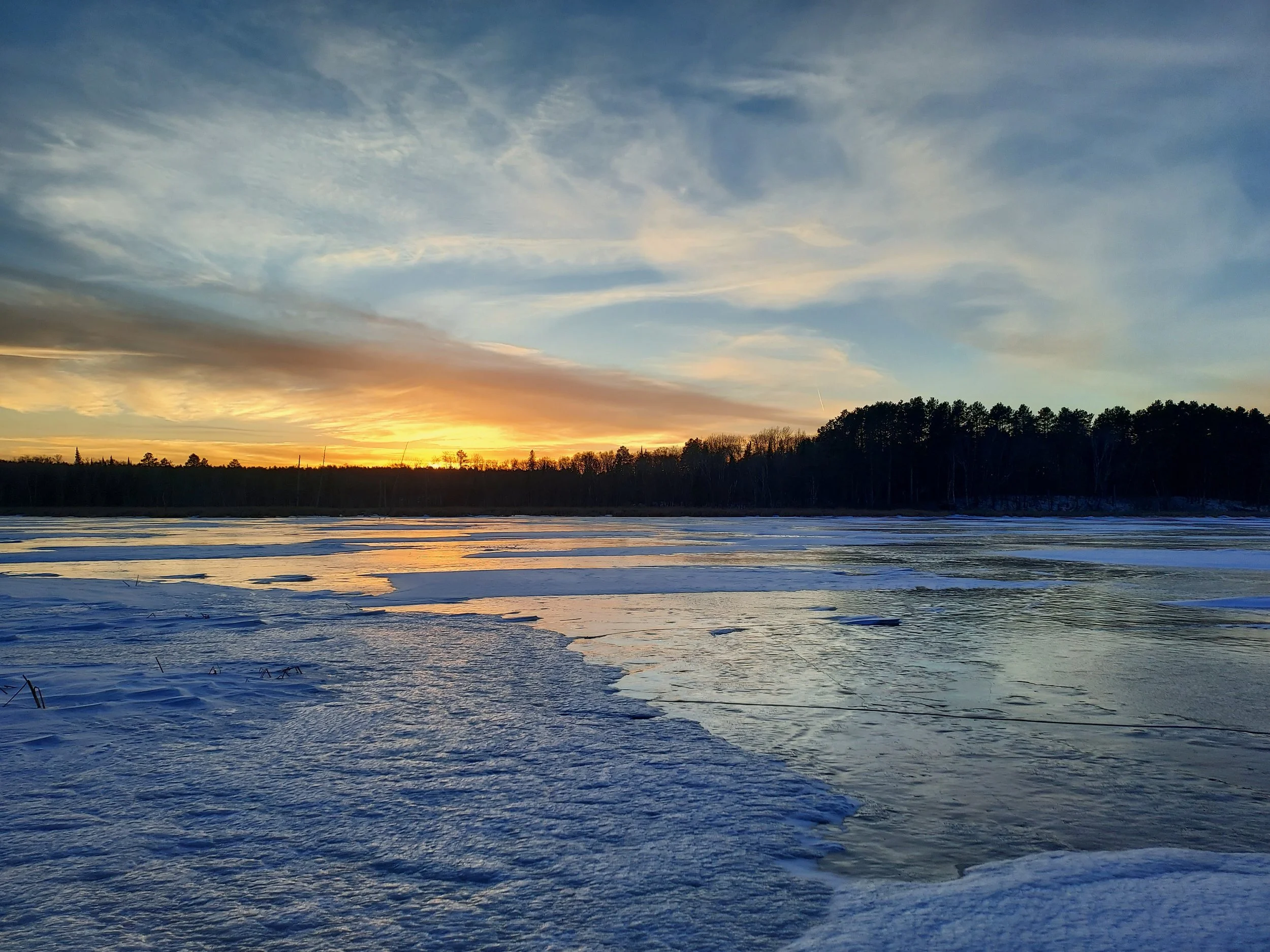 Frozen lake at sunset with snow-covered banks and trees in the distance.