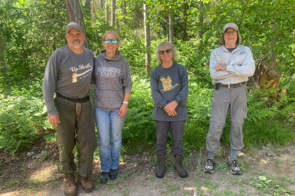 Four people standing outdoors in a wooded area, smiling at the camera. They are dressed in casual outdoor clothing.
