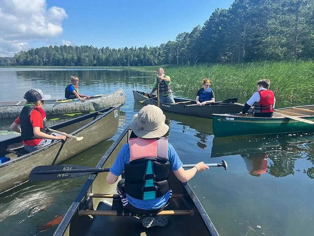 People in canoes and kayaks on a calm lake with green trees and reeds, sunny sky with a few clouds.