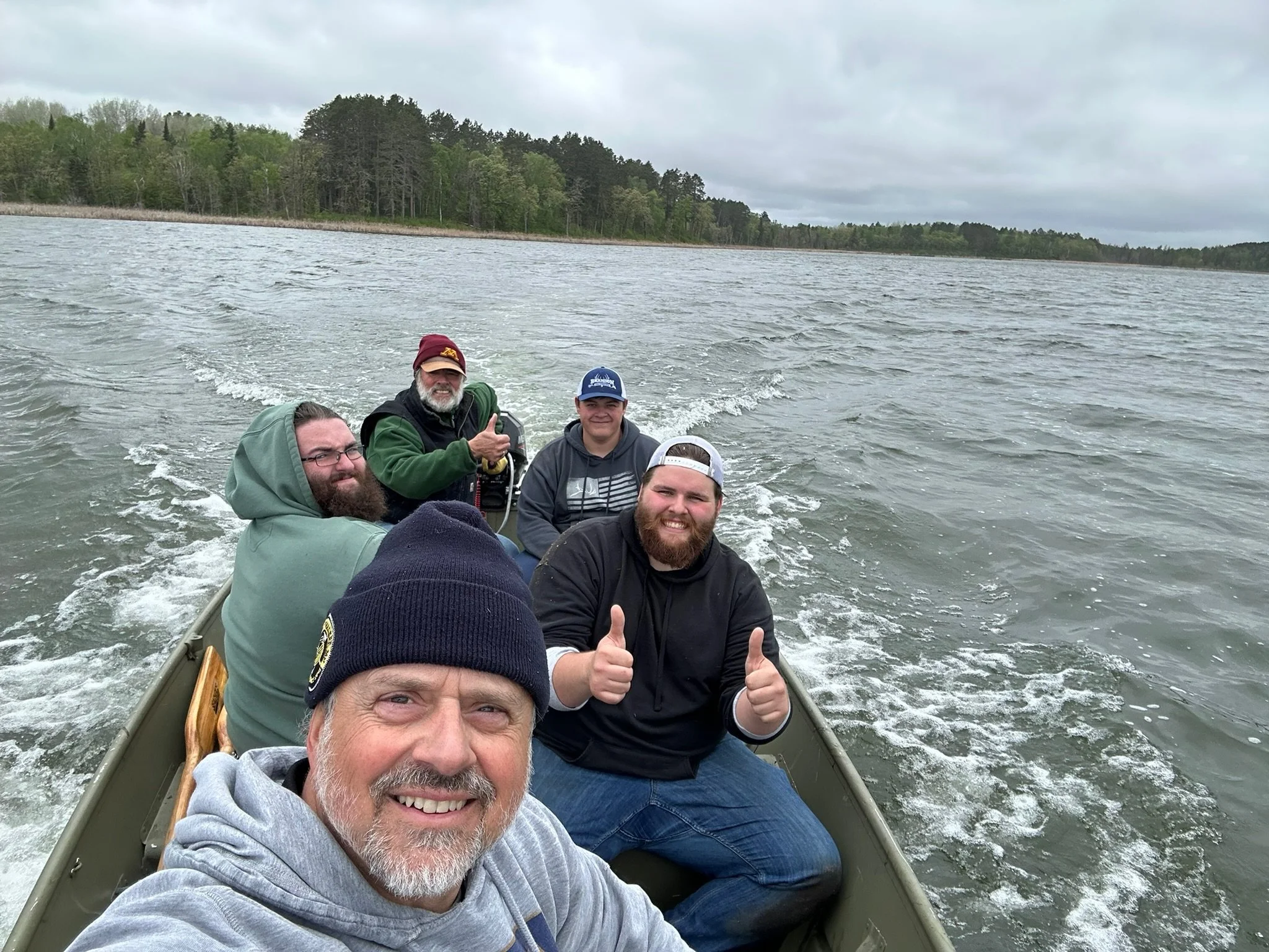 Group of five men enjoying a boat ride on a lake with a wooded shoreline in the background, some giving thumbs up and smiling.