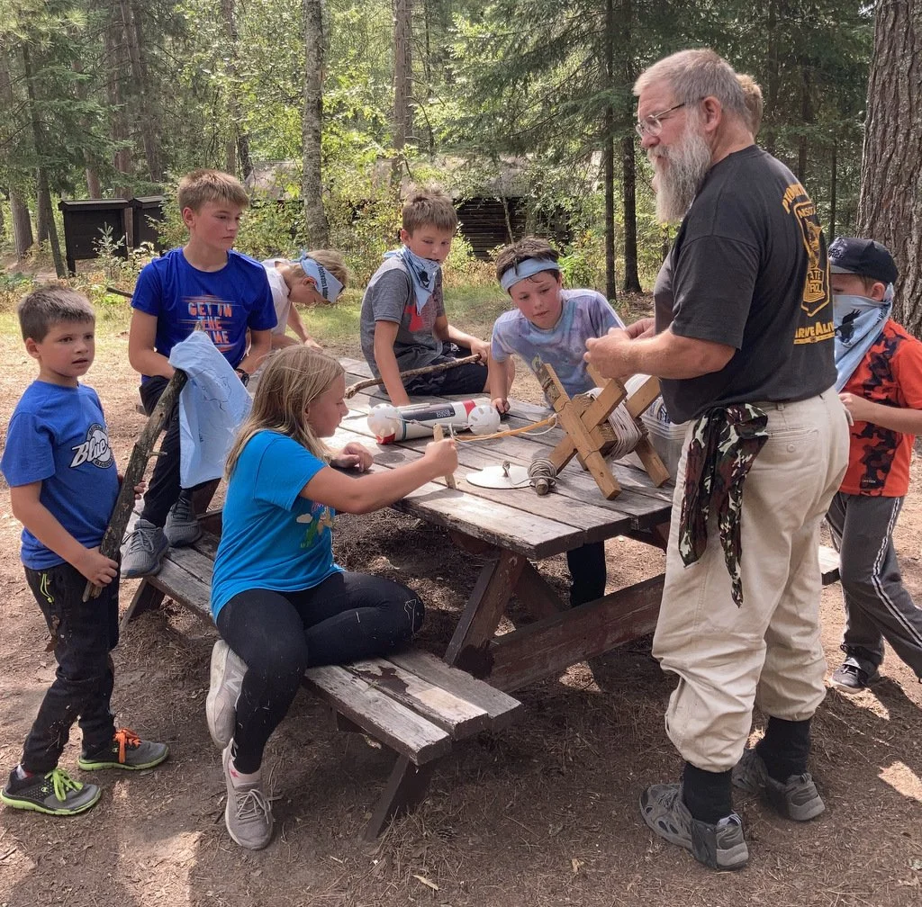 An adult man teaching a group of children about fireworks or pyrotechnics outdoors in a forest setting, with trees in the background. The children are gathered around a wooden picnic table, some sitting and some standing, observing on a learning activity.