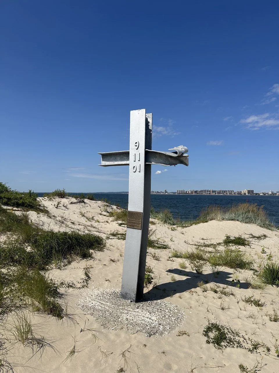 A metallic cross with a cloth draped over the horizontal beam, situated on sandy terrain with green bushes, overlooking a body of water with city buildings in the distance under a blue sky.