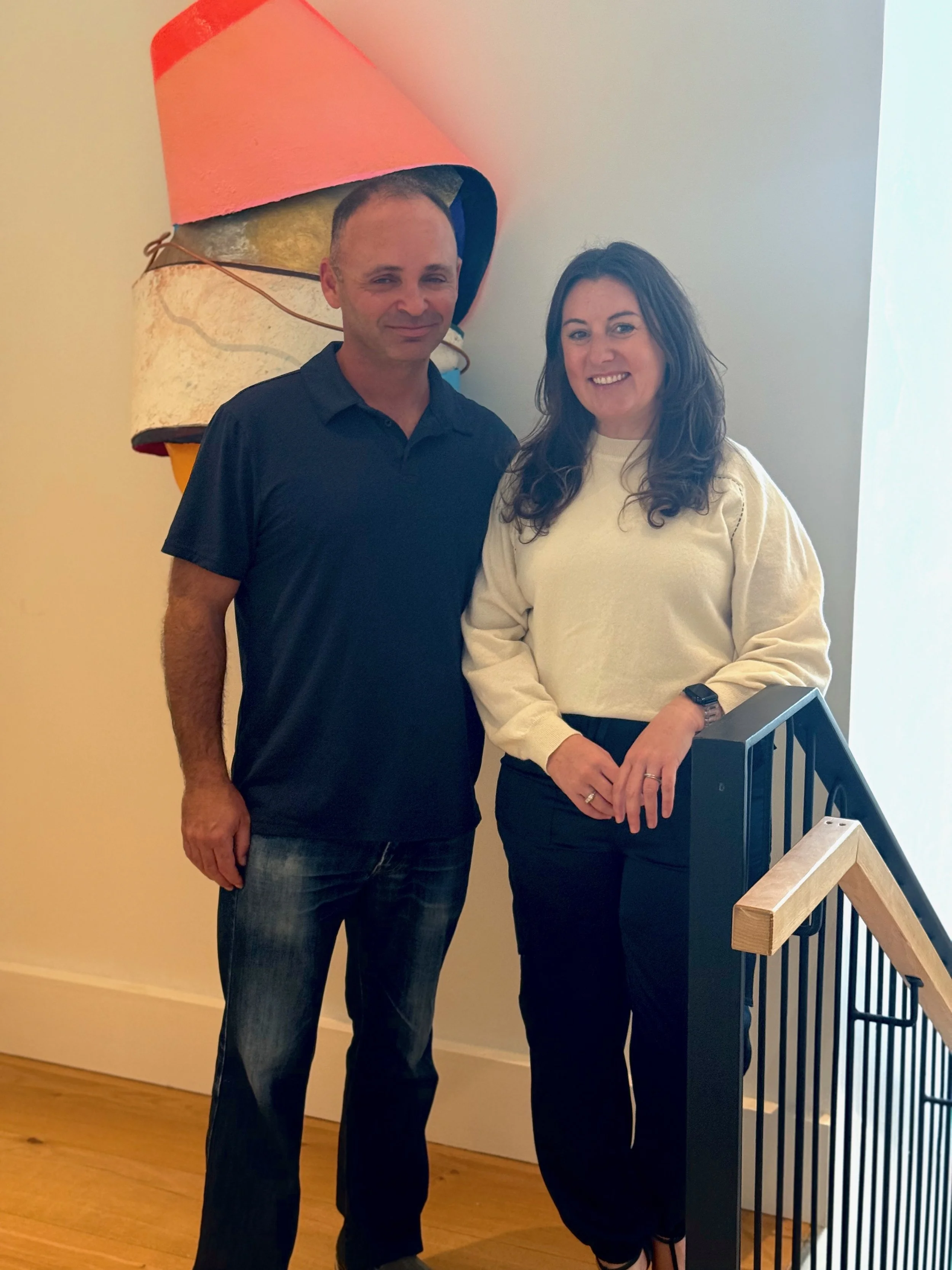 A man and woman standing indoors near a staircase with a modern railing, smiling at the camera. The man is wearing a dark polo shirt and jeans, and the woman is wearing a cream sweater and black pants.