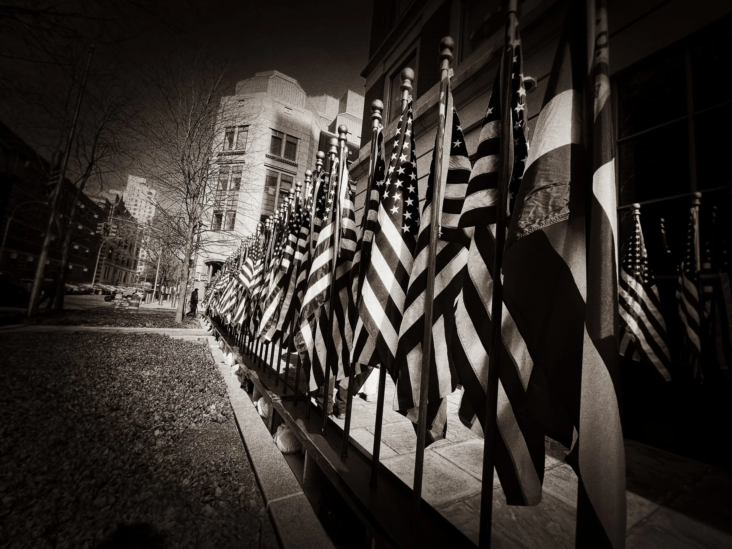 Black and white photo of American flags displayed outside a building on a city street, with trees and buildings in the background.