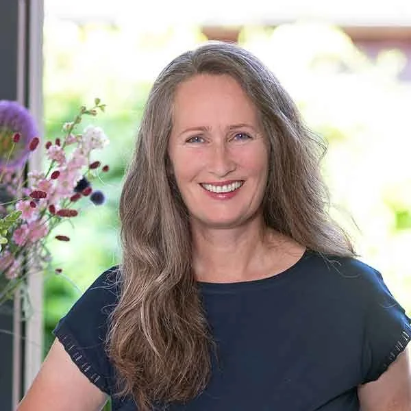 Eine lächelnde Frau mit langen, lockigen, grauen Haaren vor einem bunten Blumenstrauß.