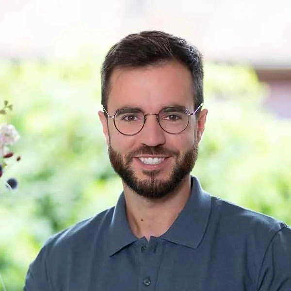 Ein Mann mit Brille und Bart, lachend, vor einem grünen, unscharfen Hintergrund, trägt eine blaue Hemd.