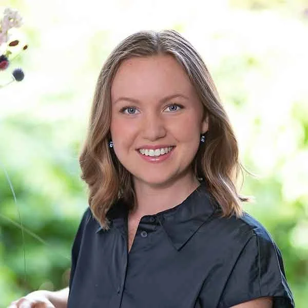 Lächelnde junge Frau mit braunen, lockigen Haaren, blauen Augen, trägt dunkle Bluse, im Hintergrund grüne Pflanzen und weißer Himmel.