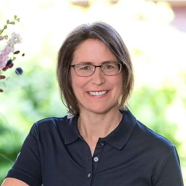 Frau mit Brille und dunkelblose Hemd, lächelnd in einem Garten.
