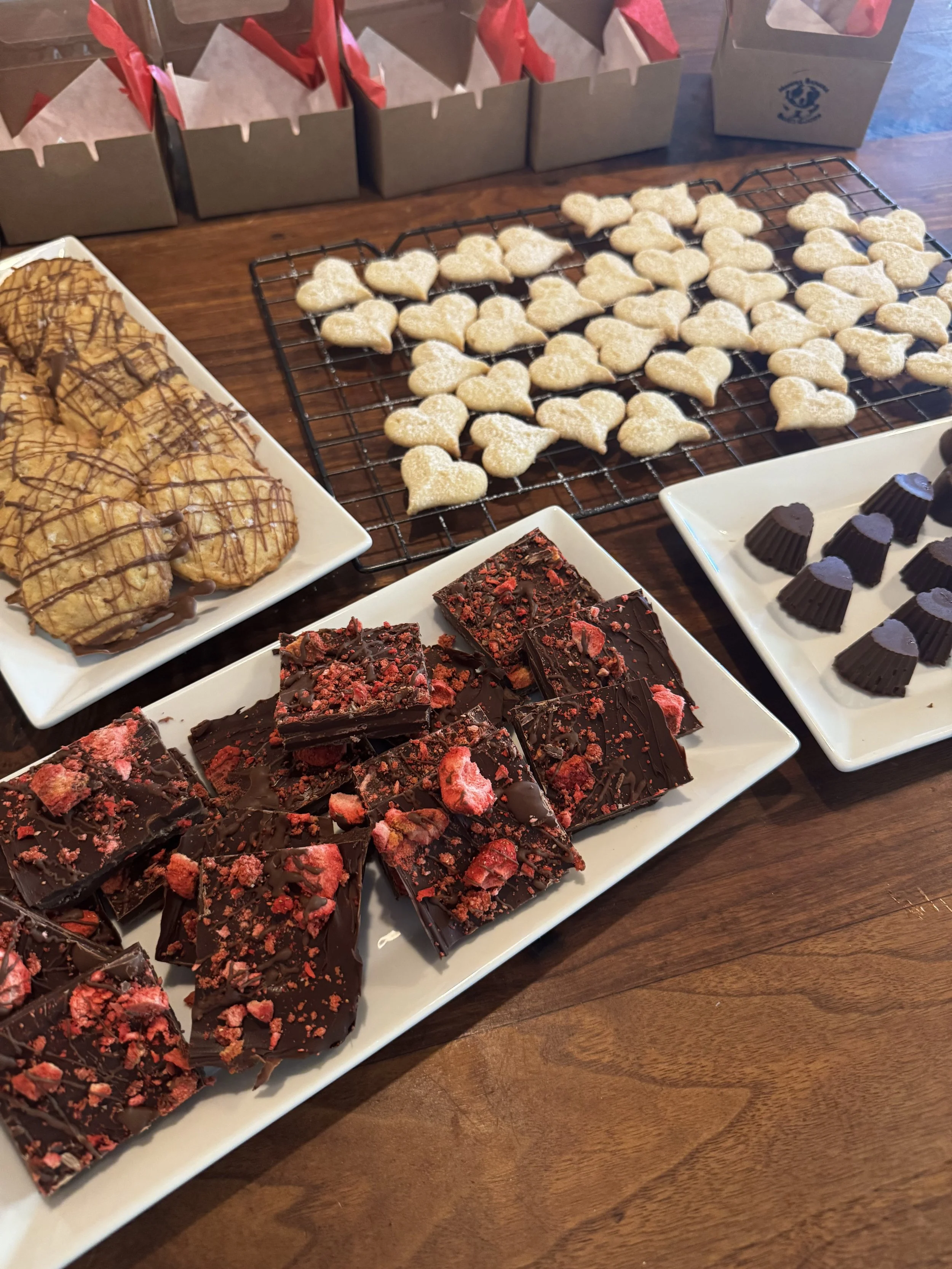 Assorted dessert treats on a table, including cookies in the shape of hearts, chocolate bark with red toppings, and chocolate cups, with gift boxes with red tissue paper in the background.
